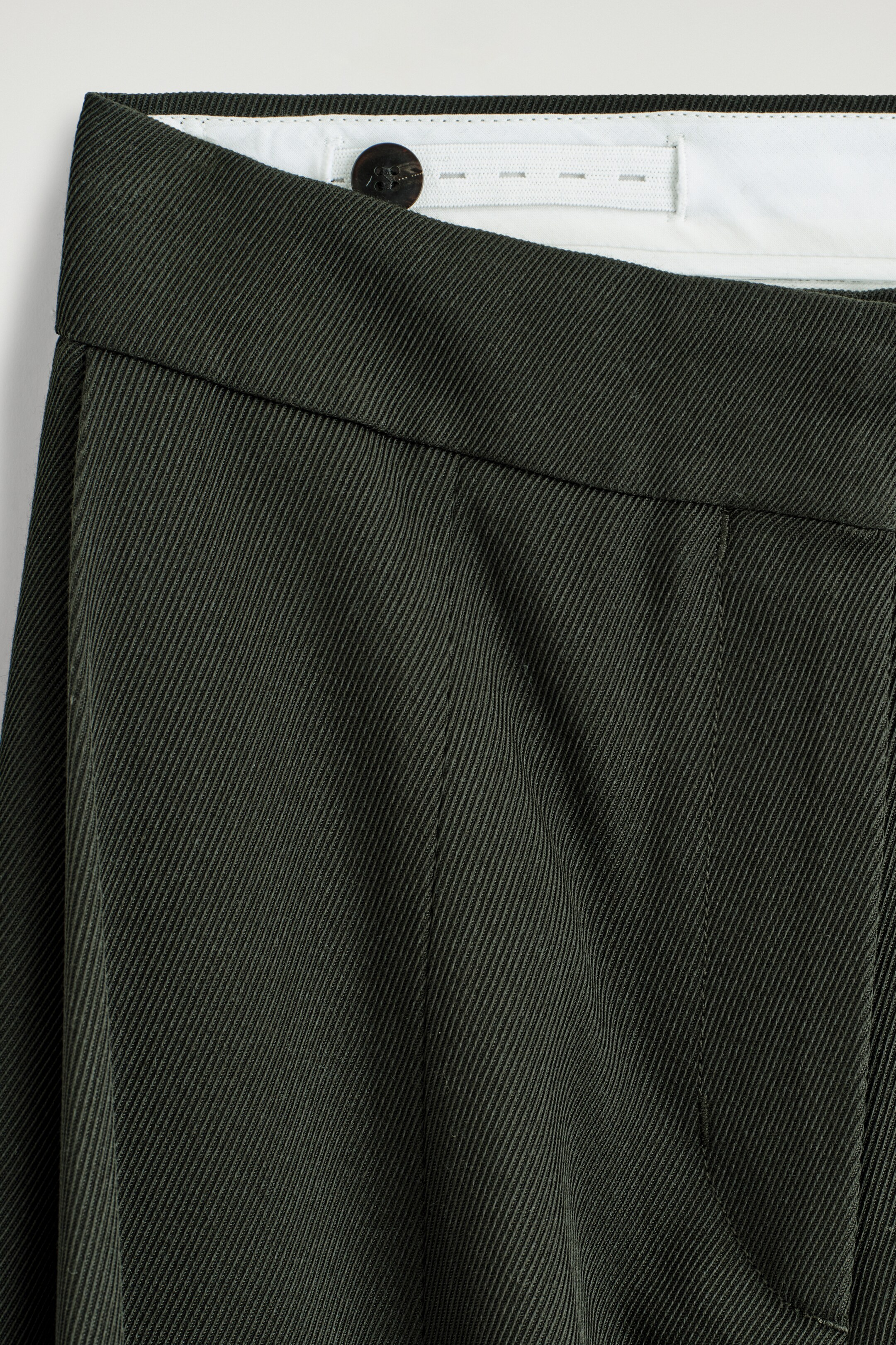 Visualizza immagine più grande: I pantaloni in twill verde scuro sono mostrati di fronte, con un primo piano sulla parte superiore della cintura. La cintura esterna pulita è visibile e il tessuto mostra un prominente intreccio diagonale del twill. È visibile anche una cintura interna regolabile con un bottone scuro.
