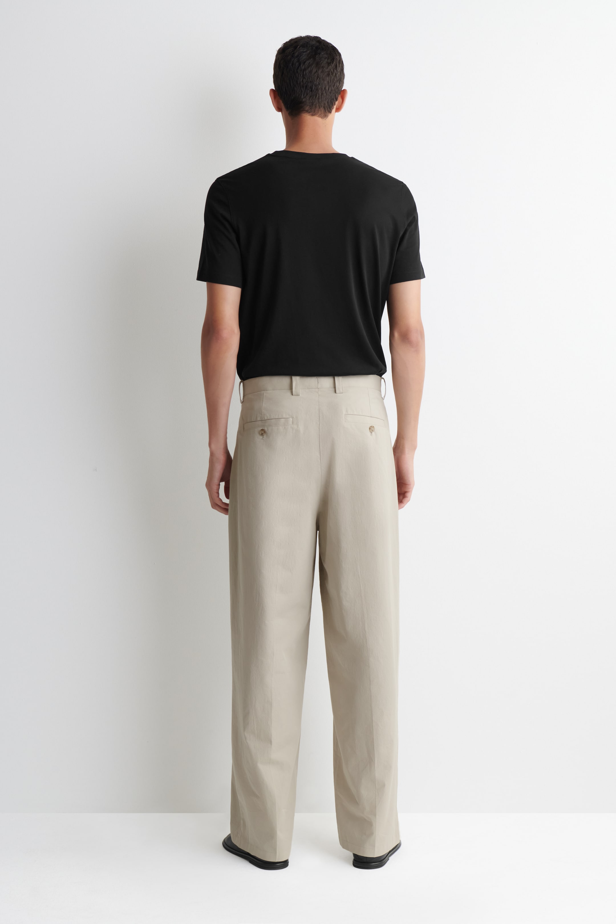 Grotere afbeelding bekijken: Een zwart T-shirt met korte mouwen en een ronde hals wordt gedragen in een lichtbeige broek met wijde pijpen, die voorzien is van riemlussen en twee achterzakken met knopen, waarbij beide kledingstukken van achteren worden bekeken.