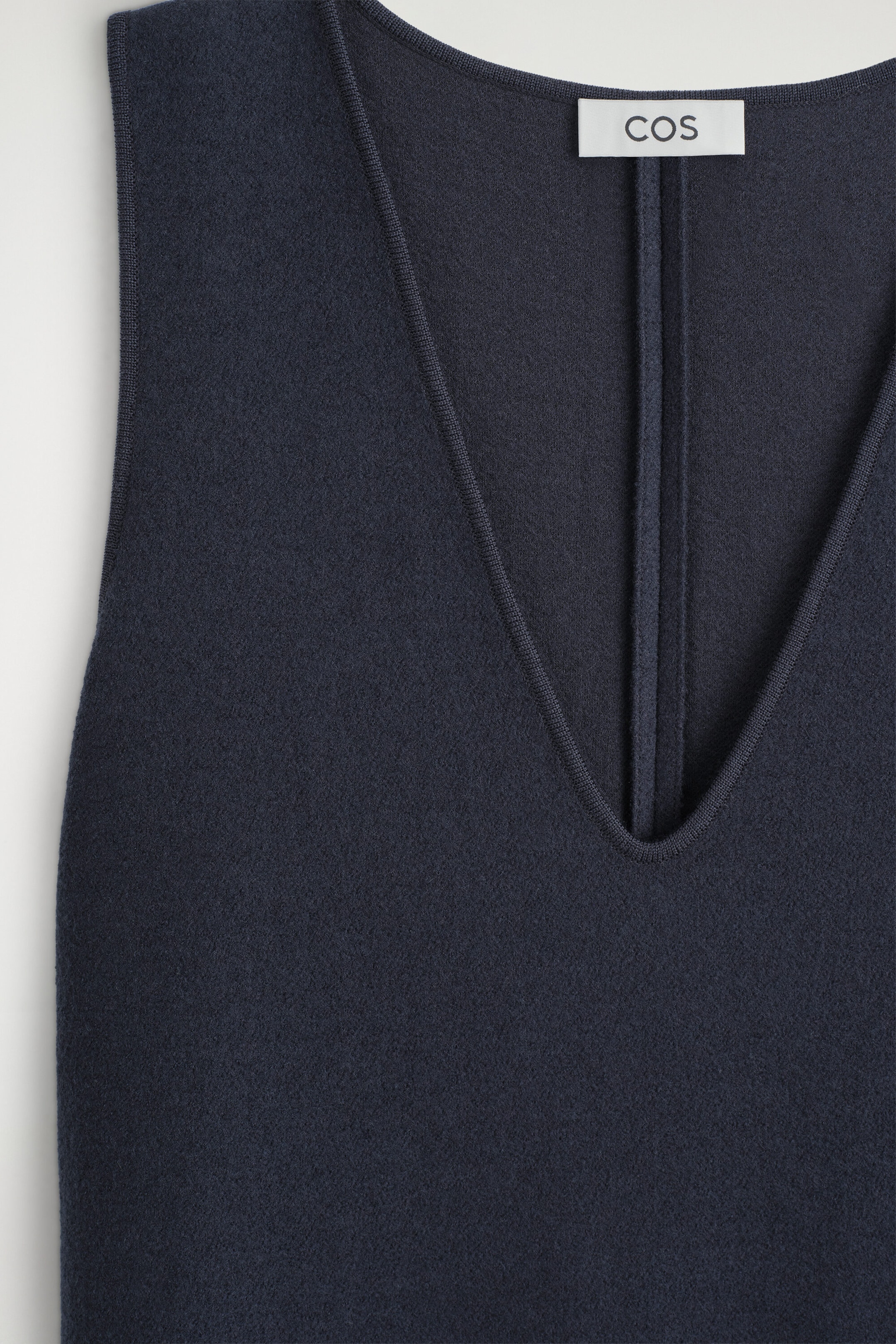 A close-up front view of the boiled wool-blend V-neck midi dress showing its deep V-neckline finished with fine ribbed trim. The sleeveless garment showcases a textured dark navy wool-blend fabric and a white brand label at the back neckline. A close-up front view of the boiled wool-blend V-neck midi dress showing its deep V-neckline finished with fine ribbed trim. The sleeveless garment showcases a textured dark navy wool-blend fabric and a white brand label at the back neckline.