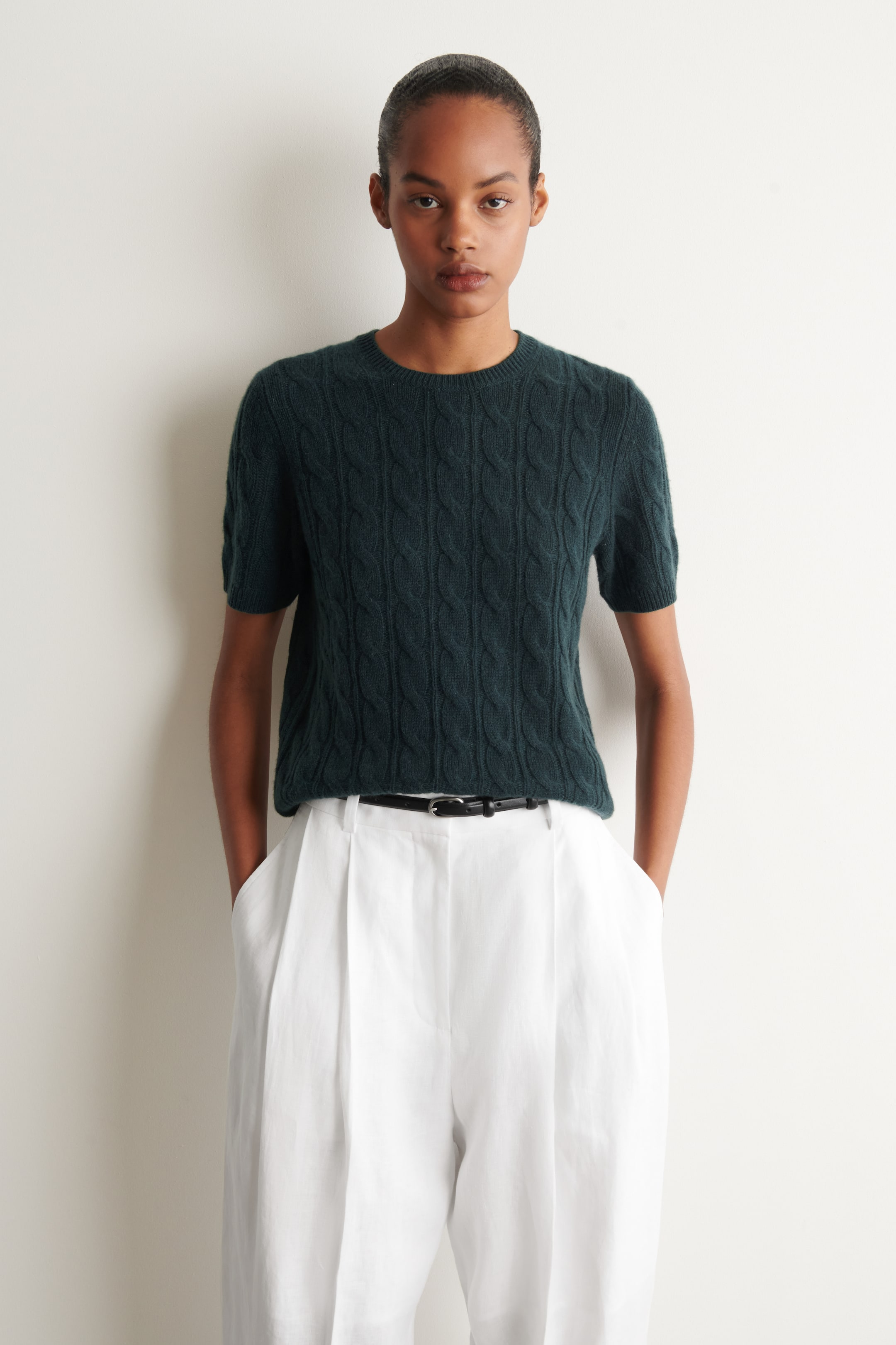 View larger image: A dark green cable-knit short-sleeve top with a ribbed crew neck, cuffs, and hem is being worn by the model, styled with white pleated trousers and a black belt.