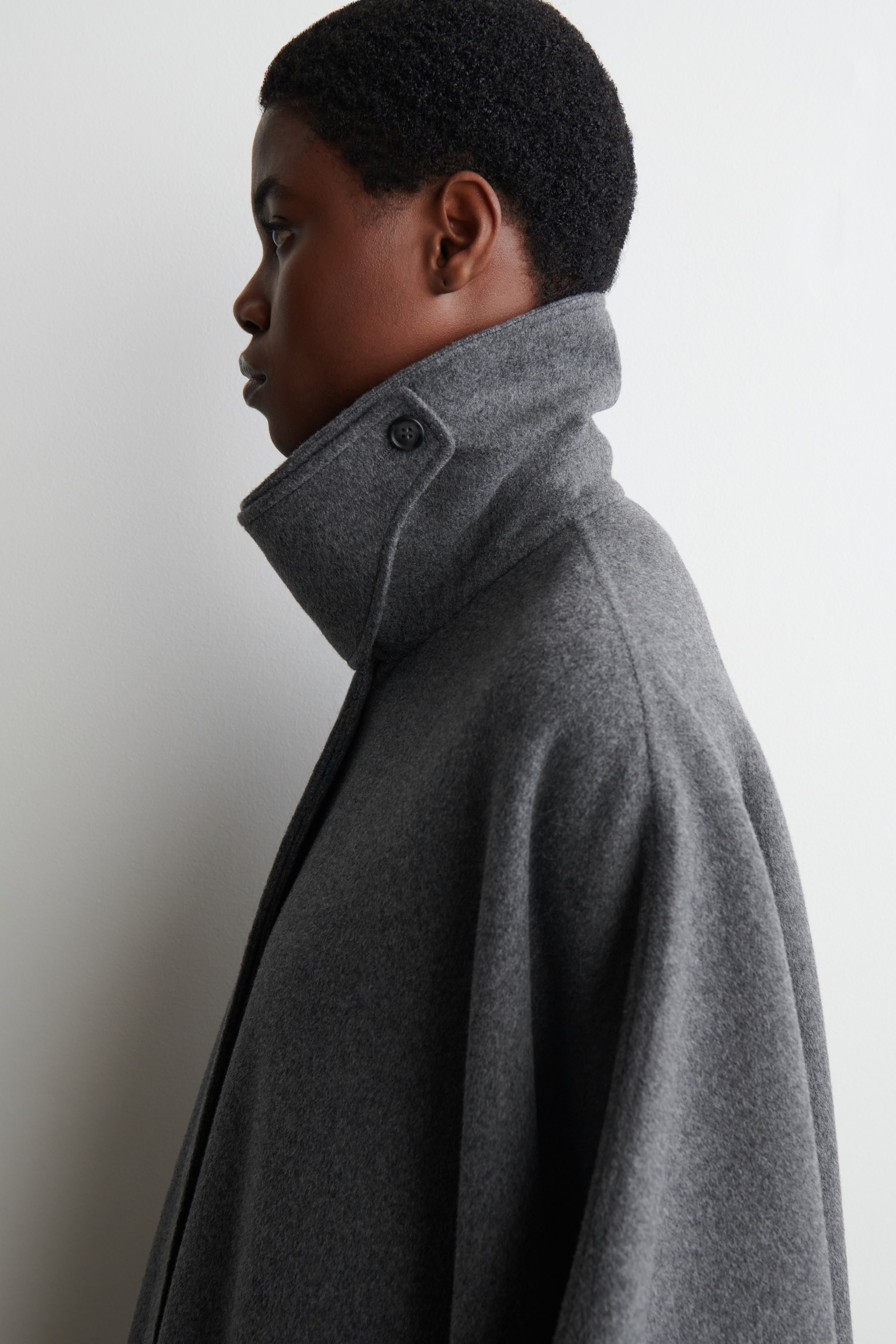 Agrandir l'image: La cape en mélange de laine grise est présentée de profil, avec son col haut montant entourant le cou. Le col présente une fermeture visible avec un bouton foncé, et le tissu de la cape drape volumineusement sur les épaules.