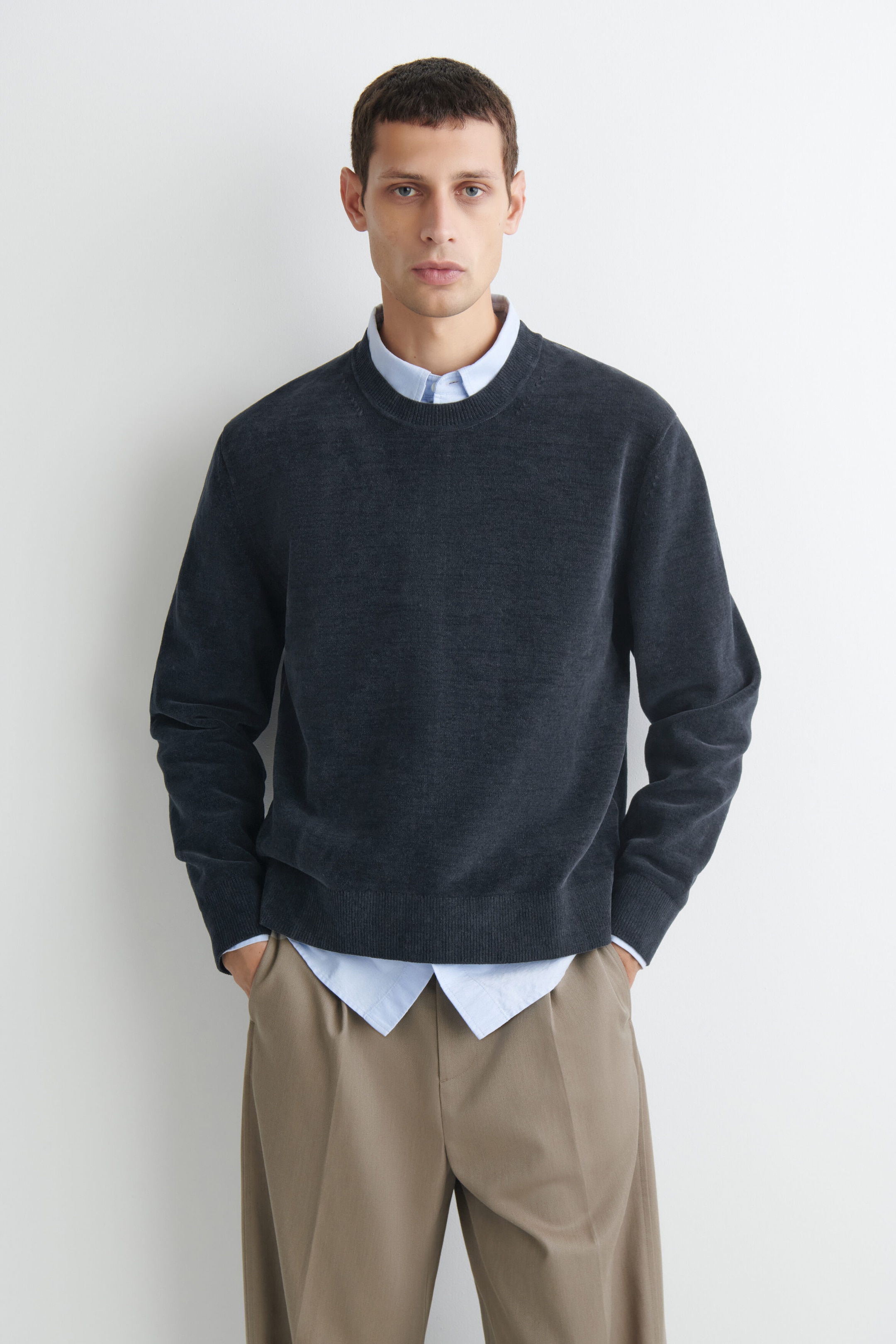The dark gray cotton-chenille crew-neck sweater is shown from the front, featuring a soft, textured finish with a ribbed neckline, cuffs, and hem. It is layered over a light blue collared shirt and styled with beige trousers. The dark gray cotton-chenille crew-neck sweater is shown from the front, featuring a soft, textured finish with a ribbed neckline, cuffs, and hem. It is layered over a light blue collared shirt and styled with beige trousers.