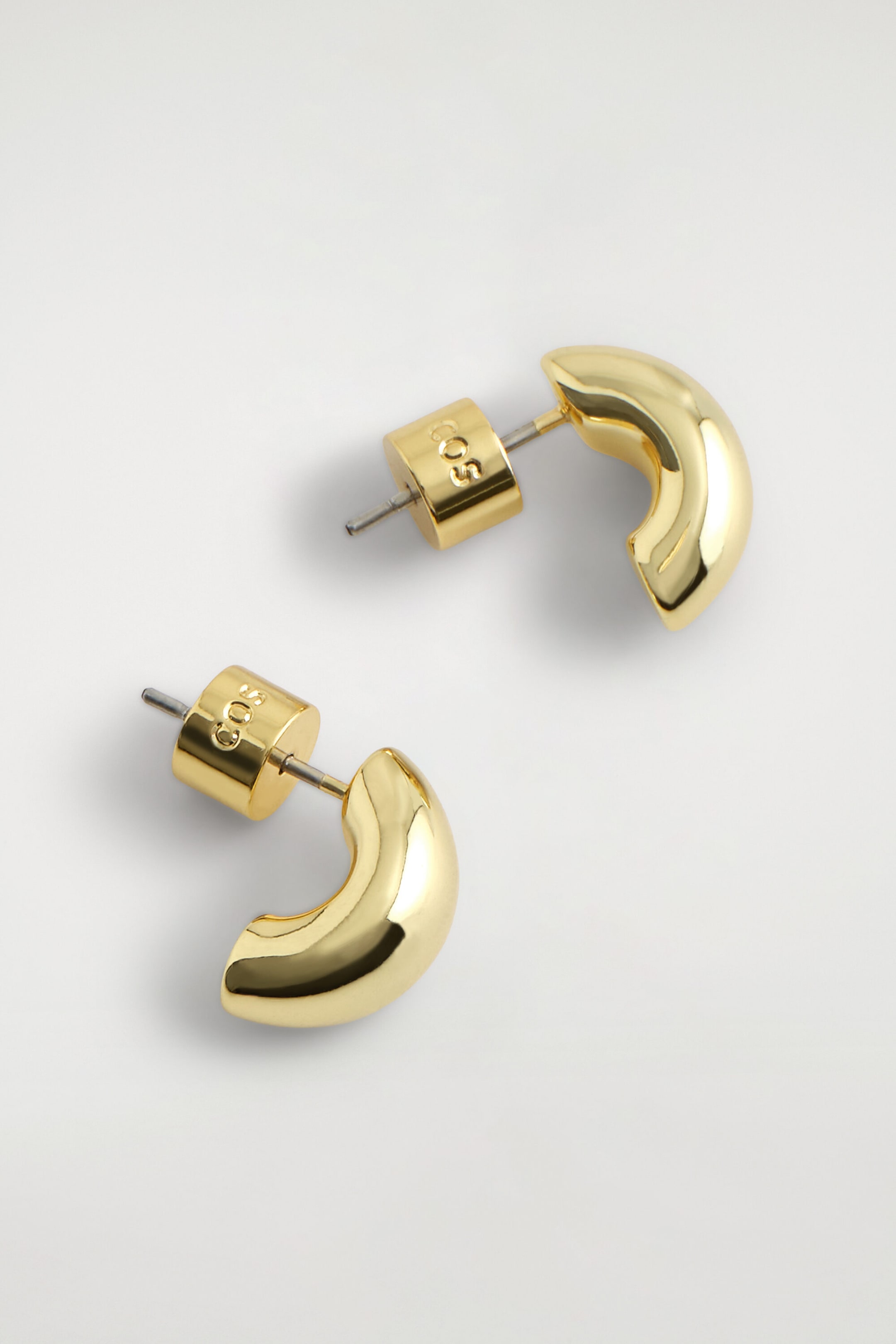Les boucles d’oreilles demi-créoles sculpturales présentent une silhouette arrondie et volumineuse, avec une finition dorée polie. Elles sont dotées de fermoirs à tige avec poussettes cylindriques gravées du logo « COS ».