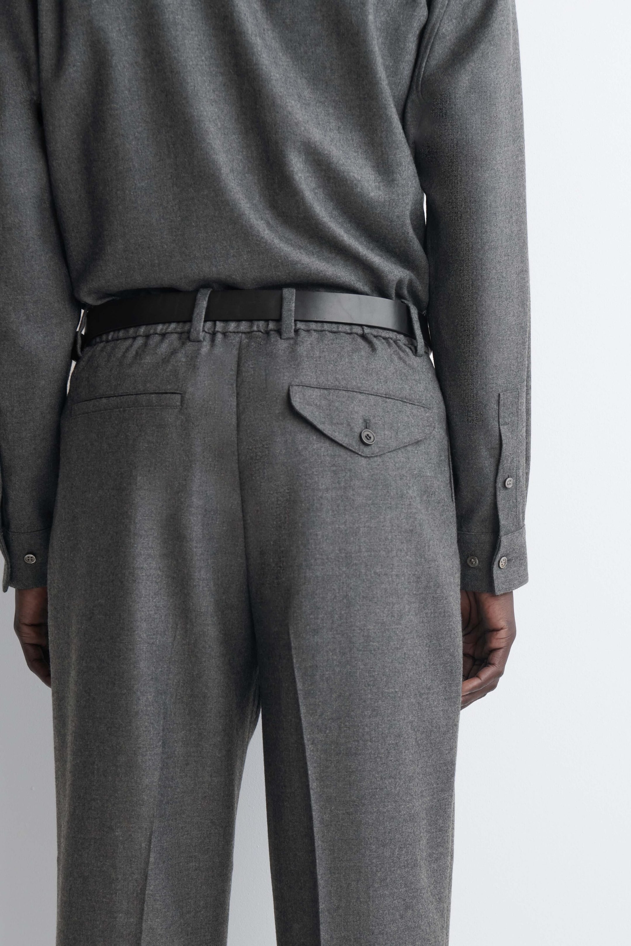 Diese grauen Hosen, von hinten gezeigt, verfügen über einen elastischen Bund mit einem schwarzen Ledergürtel und scharfen Bügelfalten entlang der Beine. Die Hose zeigt eine Paspeltasche auf der linken Seite und eine Pattentasche mit Knopf auf der rechten Seite, während ein passendes graues Langarmhemd in den Bund gesteckt ist. Diese grauen Hosen, von hinten gezeigt, verfügen über einen elastischen Bund mit einem schwarzen Ledergürtel und scharfen Bügelfalten entlang der Beine. Die Hose zeigt eine Paspeltasche auf der linken Seite und eine Pattentasche mit Knopf auf der rechten Seite, während ein passendes graues Langarmhemd in den Bund gesteckt ist.