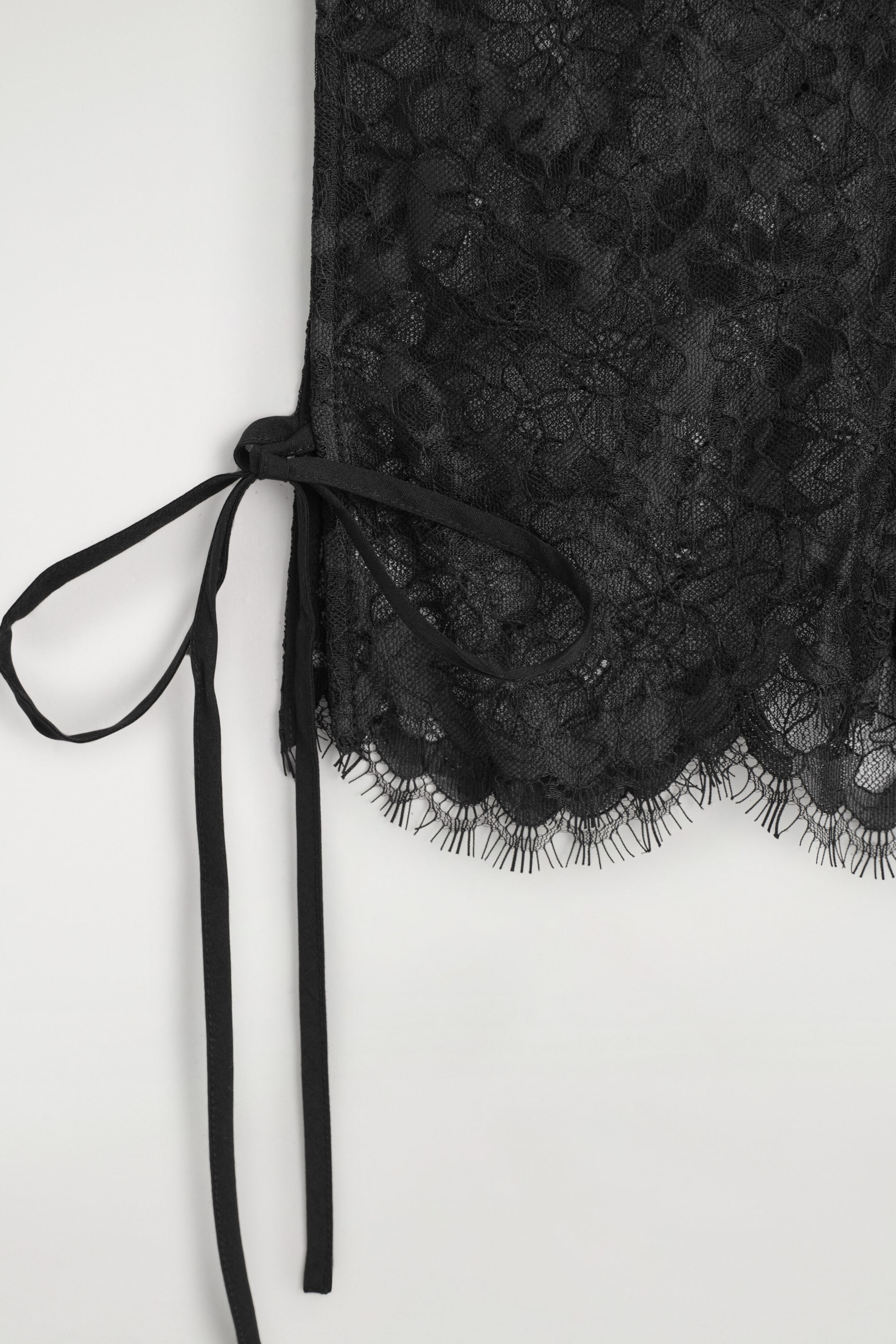 The Scalloped Lace Bib is showcasing a detailed view of its sheer black floral lace design and scalloped eyelash lace hem. A thin black fabric ribbon is tied at the side, with long straps hanging downward. The Scalloped Lace Bib is showcasing a detailed view of its sheer black floral lace design and scalloped eyelash lace hem. A thin black fabric ribbon is tied at the side, with long straps hanging downward.