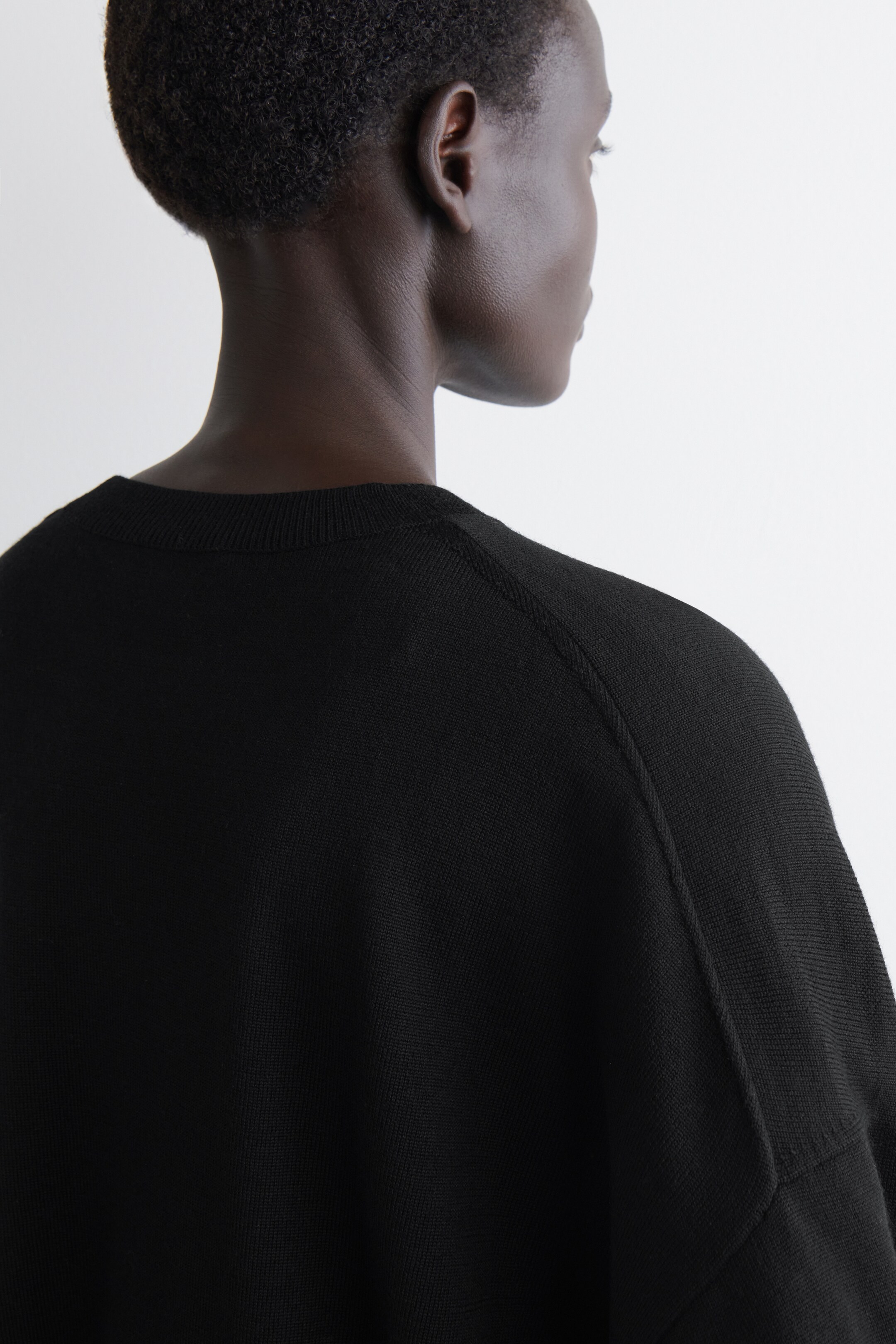 Agrandir l'image: Un pull noir en maille fine est vu de l'arrière. Il présente un col rond côtelé et des coutures raglan qui s'étendent du col le long des épaules, indiquant une coupe décontractée et surdimensionnée.