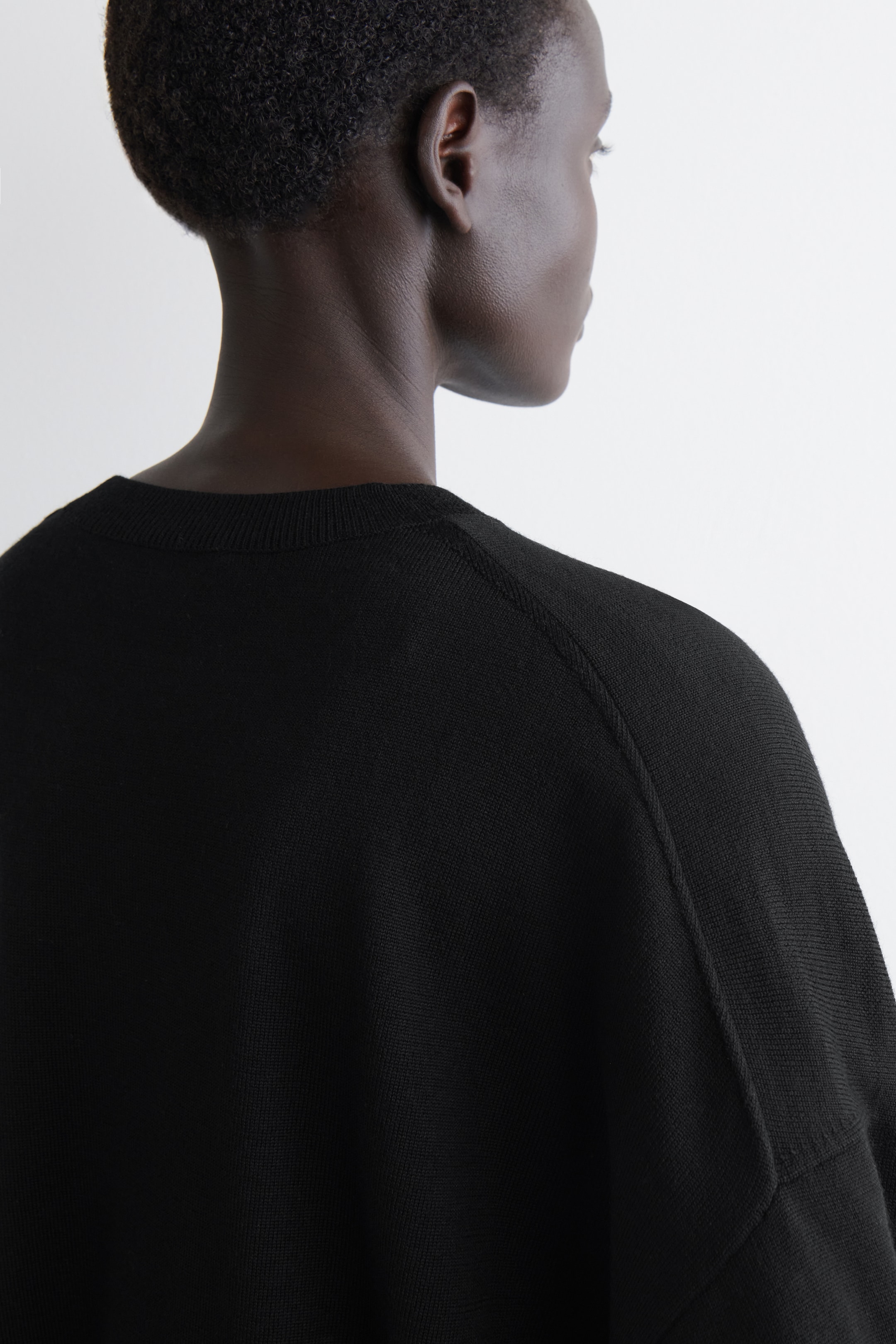 Ver imagen más grande: Un suéter negro de punto fino se está viendo desde la parte trasera. Presenta un cuello redondo acanalado y costuras raglán que se extienden desde el escote hasta los hombros, lo que indica un ajuste relajado y holgado.