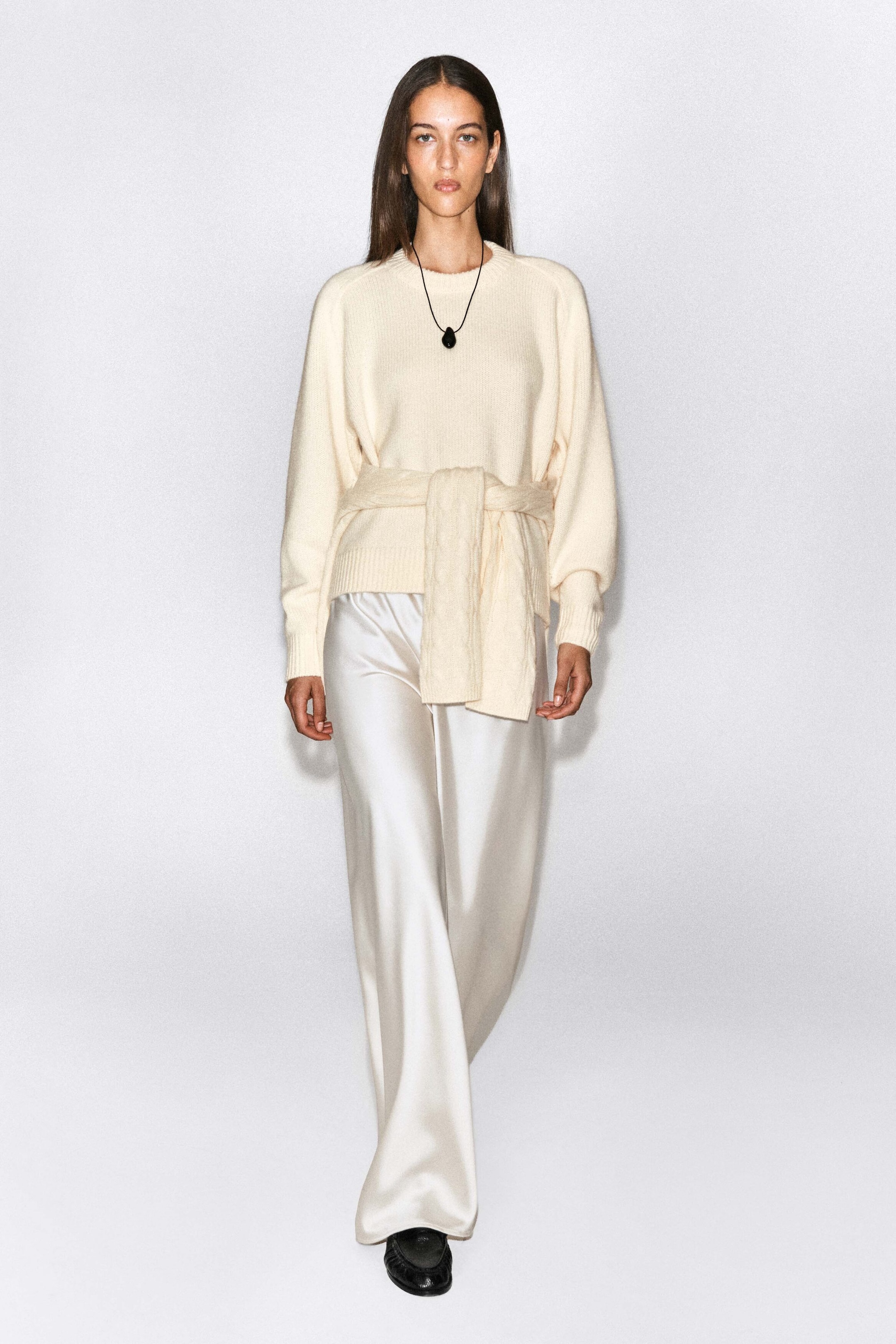 This cream-coloured rounded cashmere crew-neck jumper is being worn by the model, featuring a relaxed fit, raglan sleeves, and ribbed cuffs. A matching knit piece is tied around the waist over the jumper, which is styled with fluid, wide-leg satin trousers. This cream-coloured rounded cashmere crew-neck jumper is being worn by the model, featuring a relaxed fit, raglan sleeves, and ribbed cuffs. A matching knit piece is tied around the waist over the jumper, which is styled with fluid, wide-leg satin trousers.
