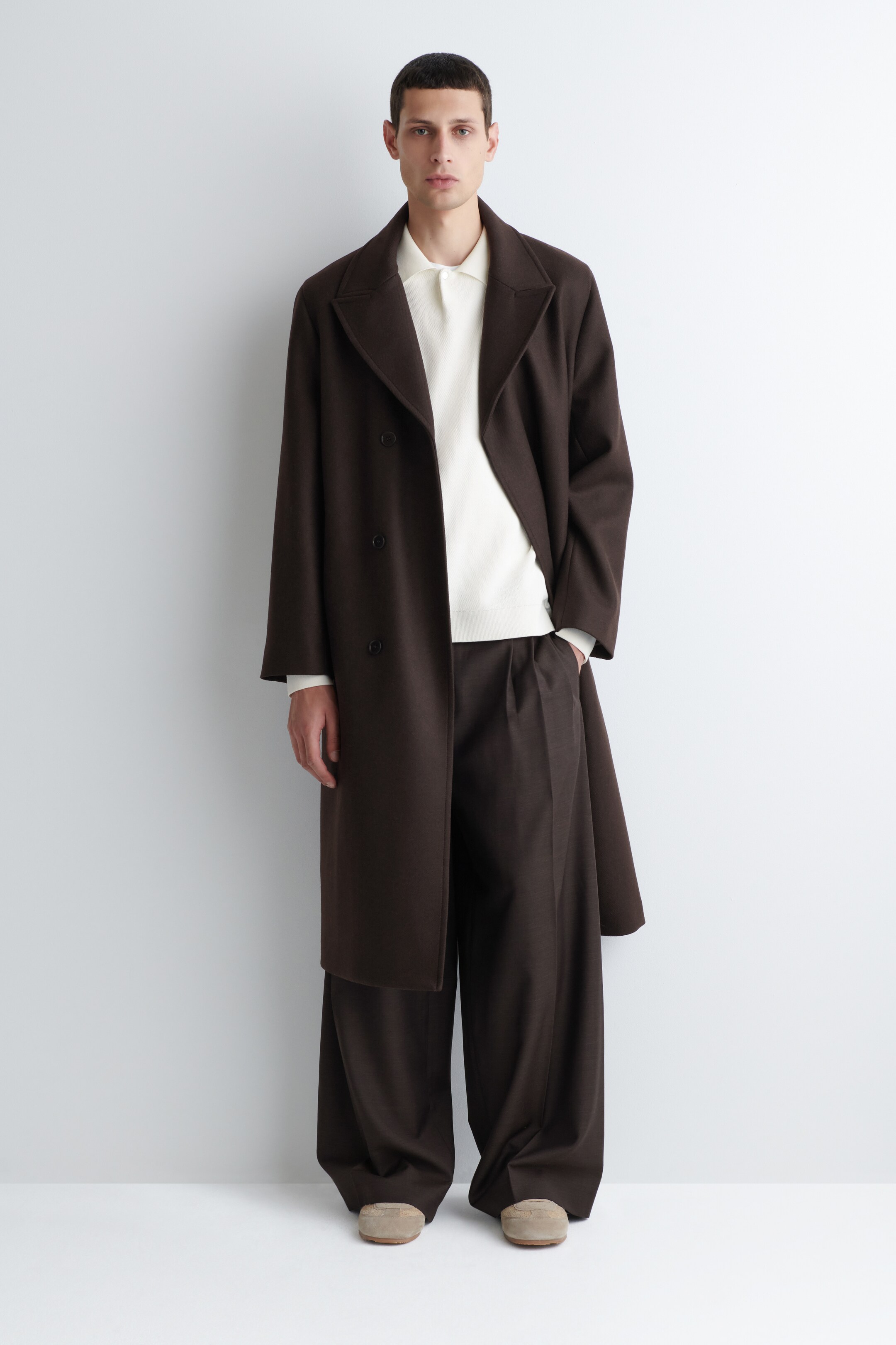 View larger image: A dark brown double-breasted coat with notch lapels and a relaxed fit is being presented in a front view, falling below the knee. It is layered over an off-white polo shirt and dark brown wide-leg trousers featuring subtle pleats, completed by tan loafers.