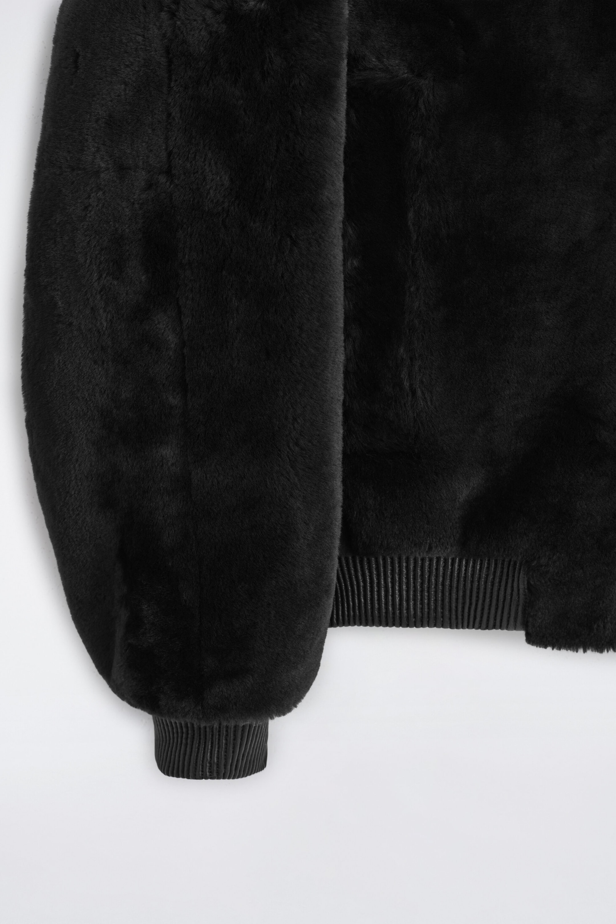 A padded shearling bomber jacket in black is shown in a close-up detail, highlighting its thick, plush shearling texture. The view emphasizes the ribbed knit cuff on the sleeve and the matching ribbed hem.