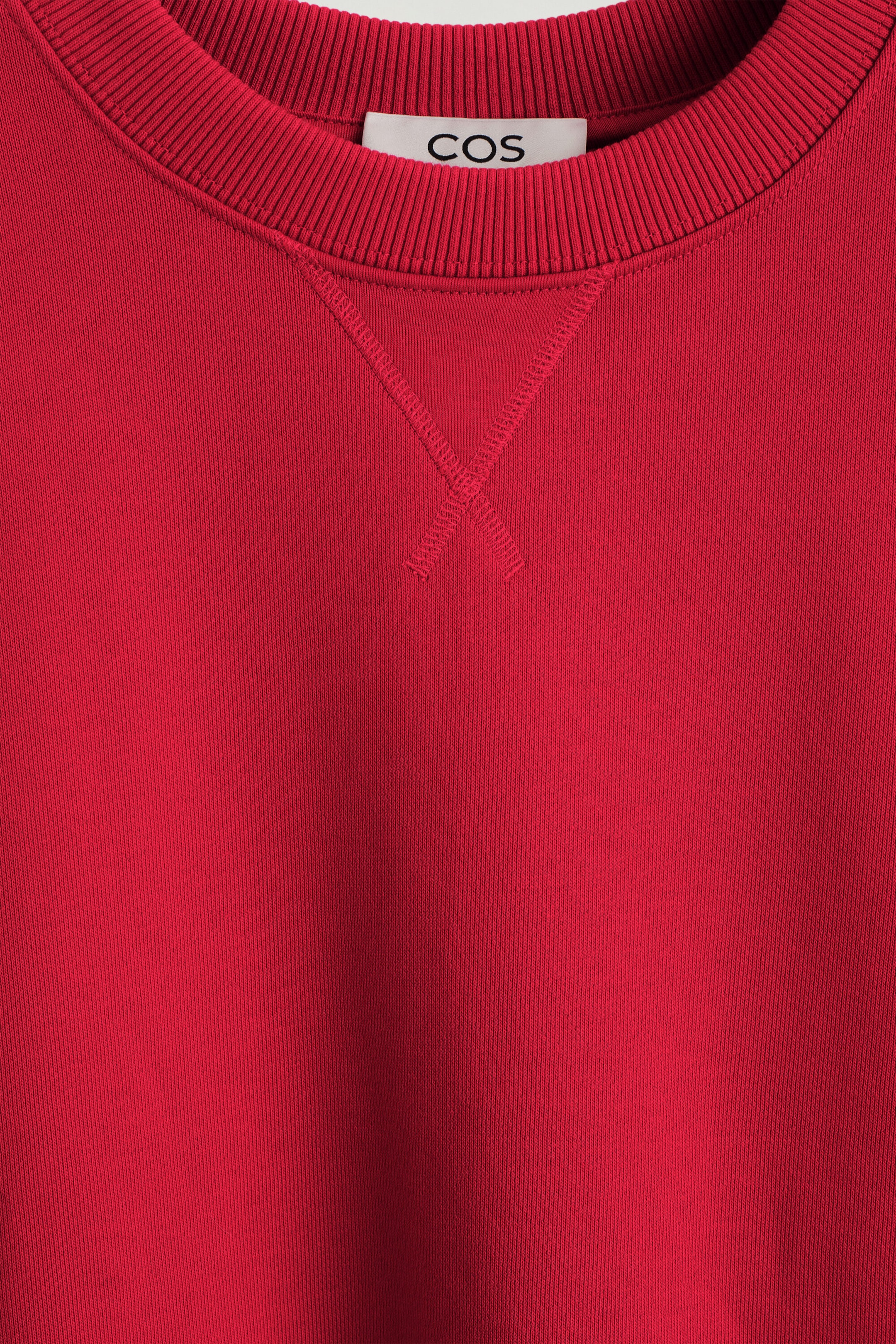 The rounded cotton crewneck sweatshirt is shown in a close-up front view, highlighting its ribbed collar and a classic triangular V-stitch detail on the upper chest. A white brand label is peeking out from the inside neckline of the red fabric.