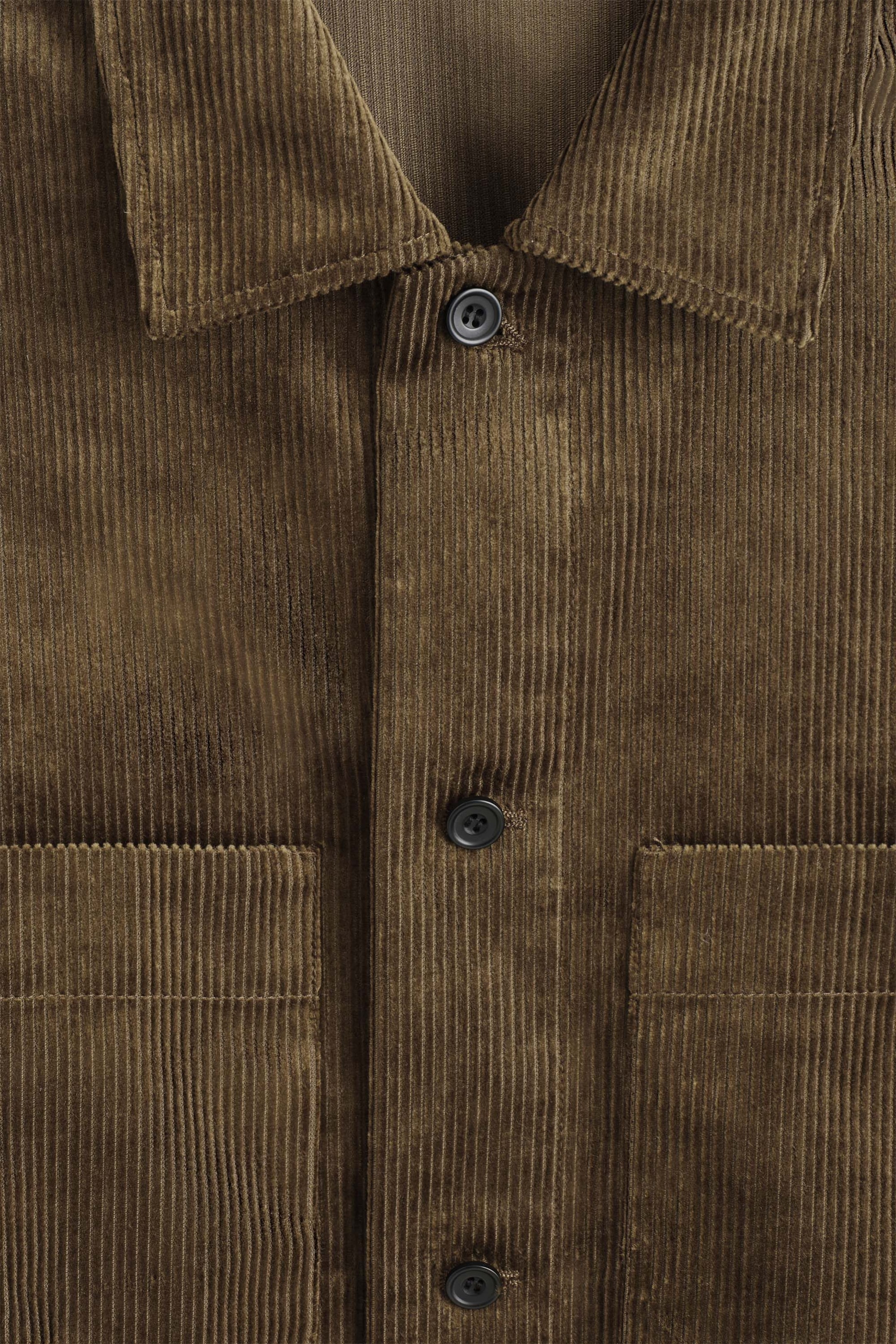 A brown cotton corduroy overshirt is shown in a close-up front view, highlighting its textured vertical ridges. The overshirt features a classic collar, a central button-down placket with dark round buttons, and two chest patch pockets. A brown cotton corduroy overshirt is shown in a close-up front view, highlighting its textured vertical ridges. The overshirt features a classic collar, a central button-down placket with dark round buttons, and two chest patch pockets.