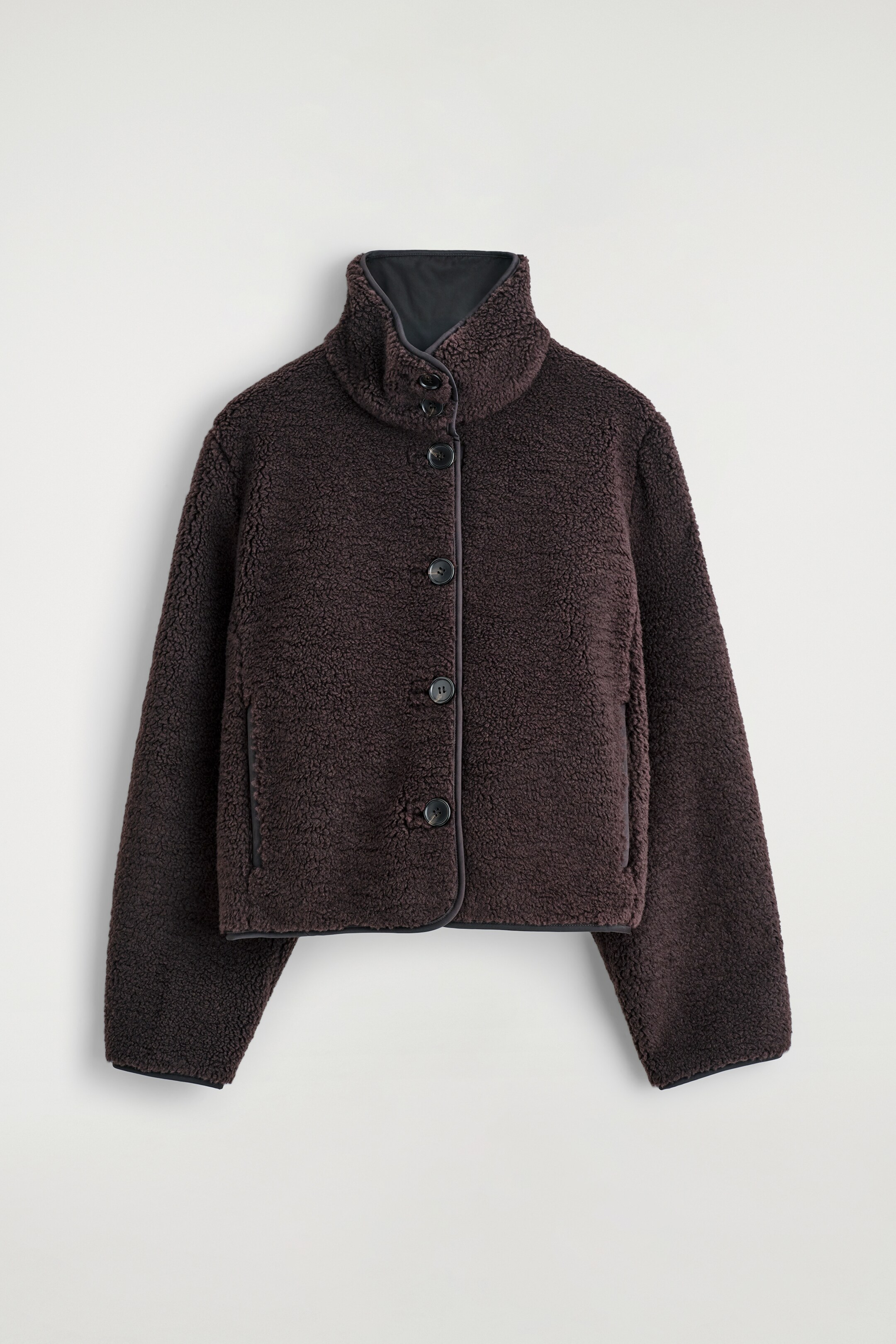 Größeres Bild ansehen: Eine dunkelbraune, strukturierte Fleecejacke zeigt ihre Vorderansicht, mit einem hohen Stehkragen mit dunklerer Fütterung, einem Druckknopfverschluss mit vier dunklen Knöpfen und dunklen kontrastierenden Abschlüssen entlang der Knopfleiste, am Kragen, Saum und an den zwei vertikalen Seitentaschen.