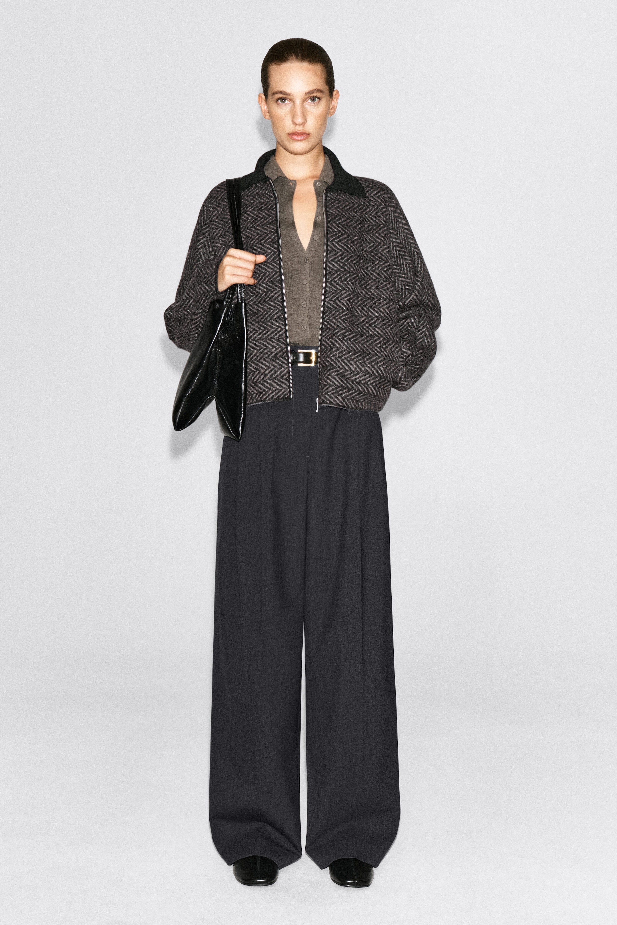 Une veste en tricot à motif chevron, de couleur gris foncé, est portée par le mannequin par-dessus un cardigan gris boutonné et un pantalon noir à jambe large. La veste présente un col côtelé noir et une fermeture éclair frontale argentée, et le mannequin tient un sac à bandoulière en cuir noir.