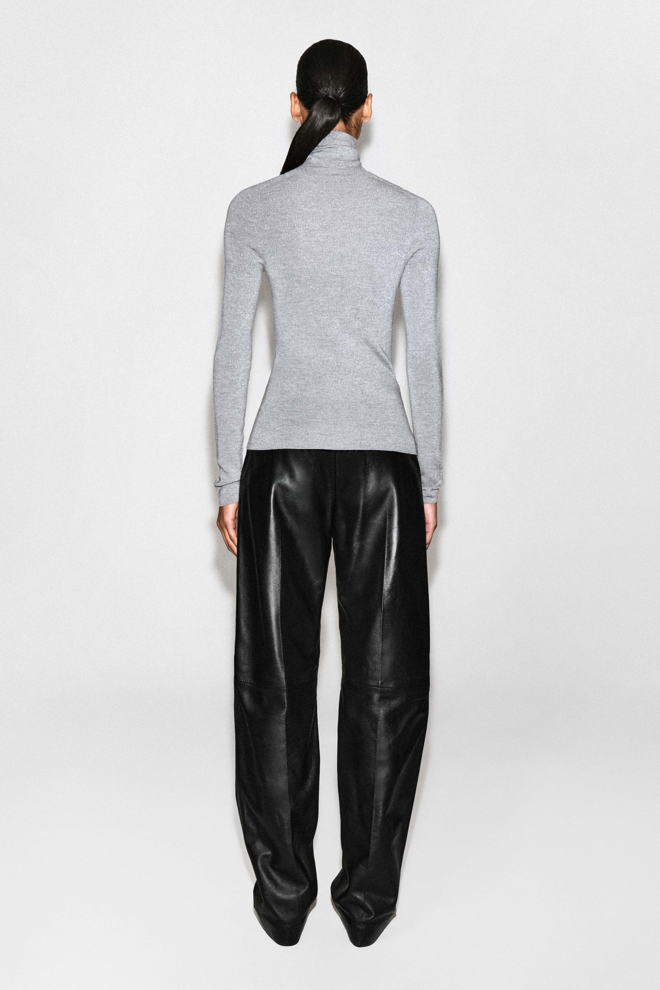 The sparkly seamless wool funnel-neck top is shown from the back, highlighting its slim-fit silhouette, long sleeves, and high funnel neck. The light gray knit top drapes smoothly over the waistband of black leather trousers, showcasing its seamless construction and clean hemline.
