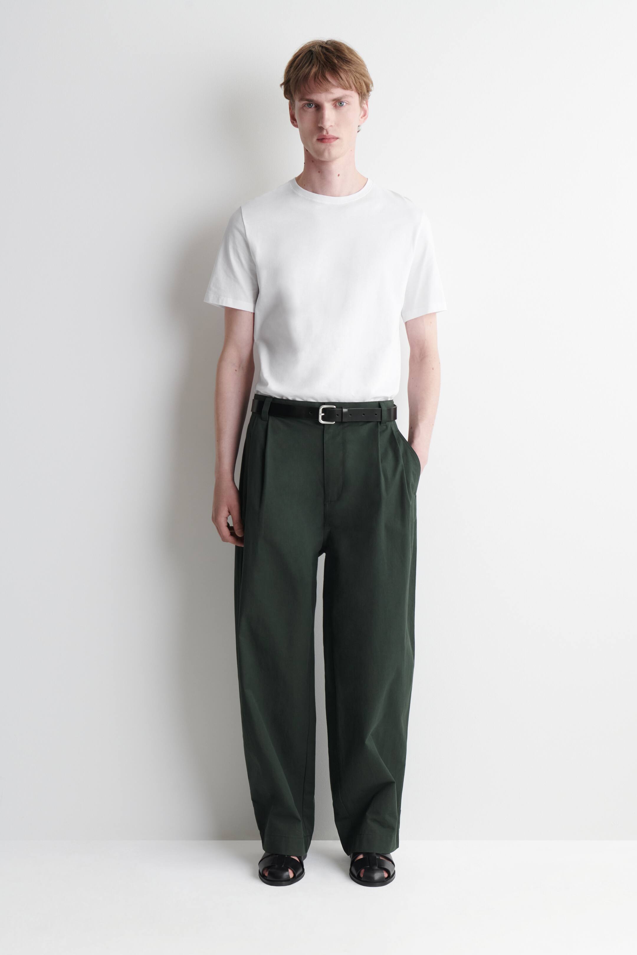 Ver imagen más grande: Una camiseta blanca de cuello redondo está metida por dentro de unos pantalones anchos plisados de color verde oscuro, ceñidos a la cintura por un cinturón negro con hebilla de tono plateado, y en los pies se llevan sandalias negras de tiras.