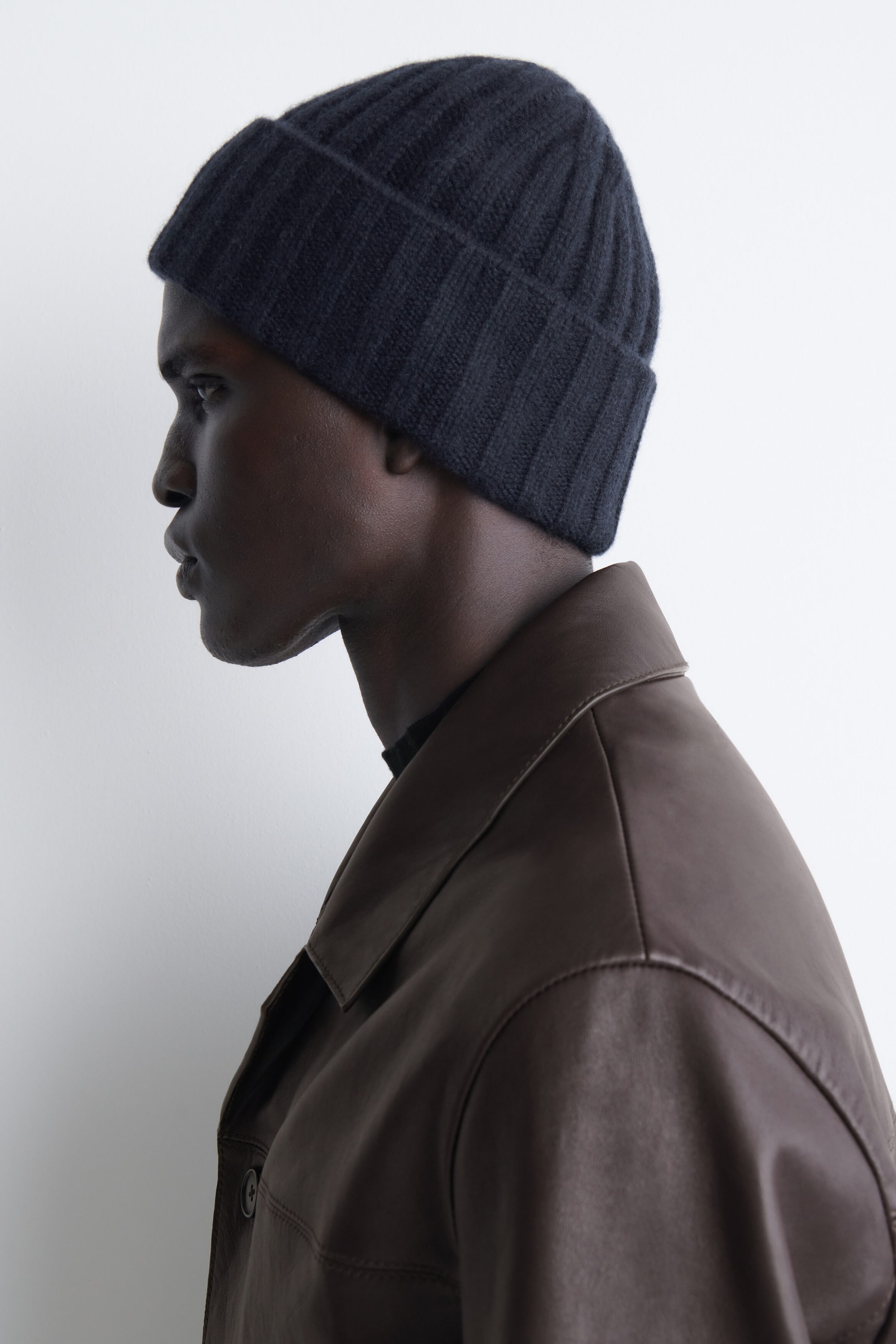 A model facing left is wearing the navy blue chunky ribbed cashmere beanie, showcasing its thick, vertical rib-knit texture and folded-over cuff. The beanie fits snugly over the head, demonstrating its structured shape and soft, heavyweight material.