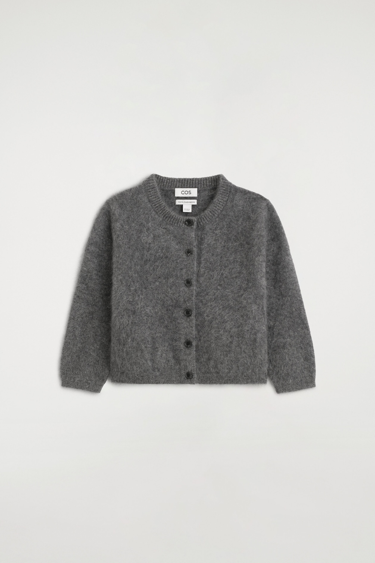 A kids' brushed pure cashmere cardigan in gray is laid flat in a front-facing view, showcasing a crew neck, button-down front, and long sleeves. The cardigan displays a soft, fuzzy texture with ribbed trim along the collar, cuffs, and hem.