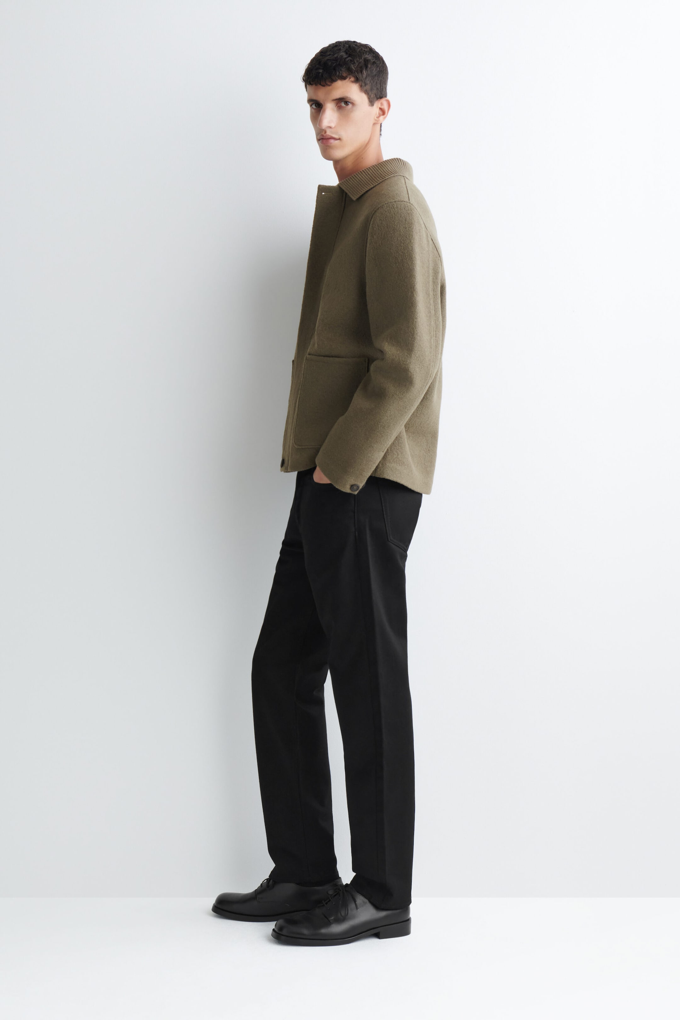A ribbed-collar boiled-wool chore jacket in olive green is worn by the model, who is standing in profile with one hand in the pocket of black trousers. The jacket features a ribbed collar, front button closure, and large patch pockets, styled with black leather shoes. A ribbed-collar boiled-wool chore jacket in olive green is worn by the model, who is standing in profile with one hand in the pocket of black trousers. The jacket features a ribbed collar, front button closure, and large patch pockets, styled with black leather shoes.