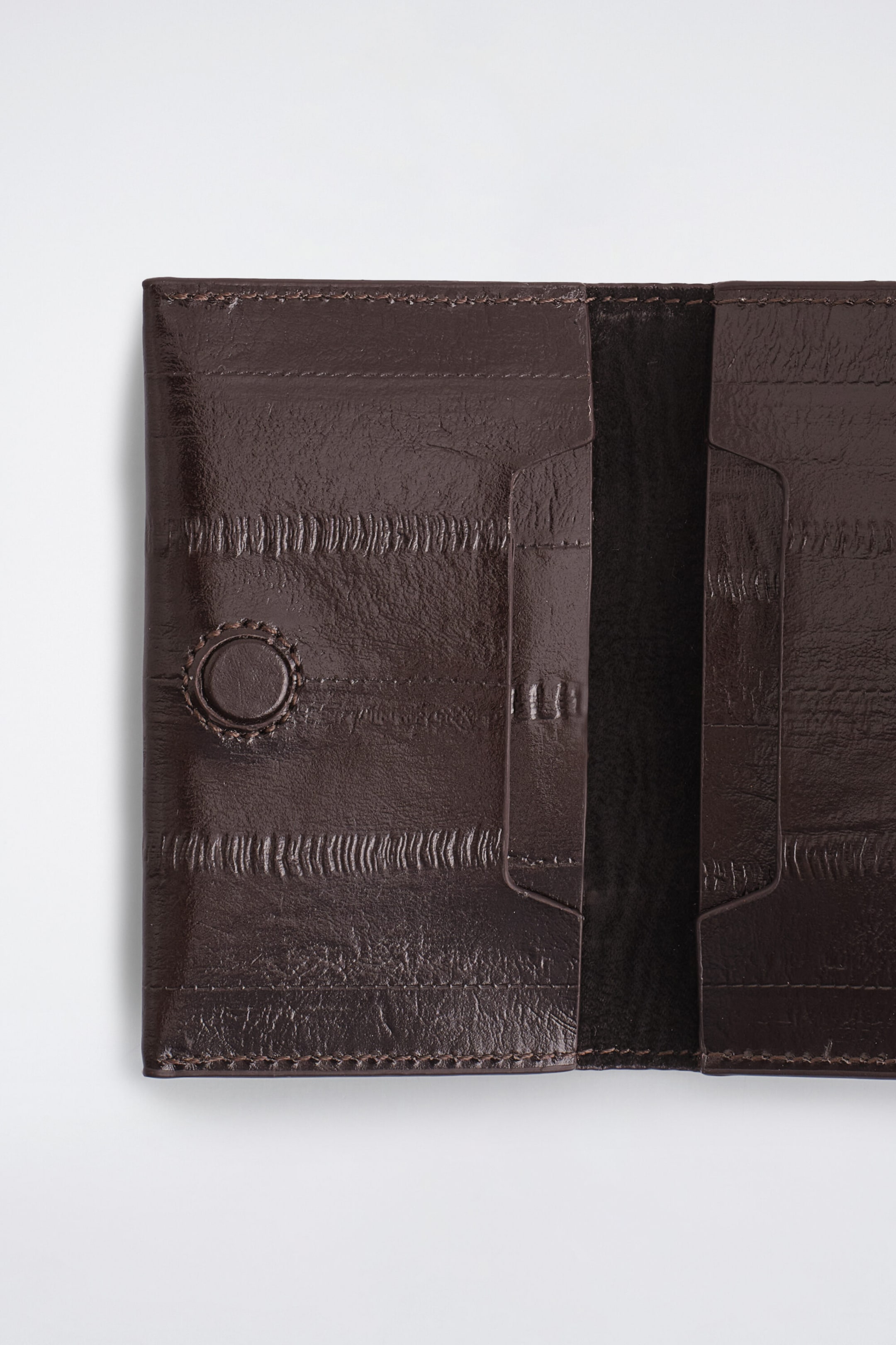 A dark brown eel-effect leather cardholder lies open, showing its interior compartments with card slots on both sides. The glossy, textured leather features distinctive striped paneling, tonal stitching, and a leather-covered snap fastener on the left panel. A dark brown eel-effect leather cardholder lies open, showing its interior compartments with card slots on both sides. The glossy, textured leather features distinctive striped paneling, tonal stitching, and a leather-covered snap fastener on the left panel.