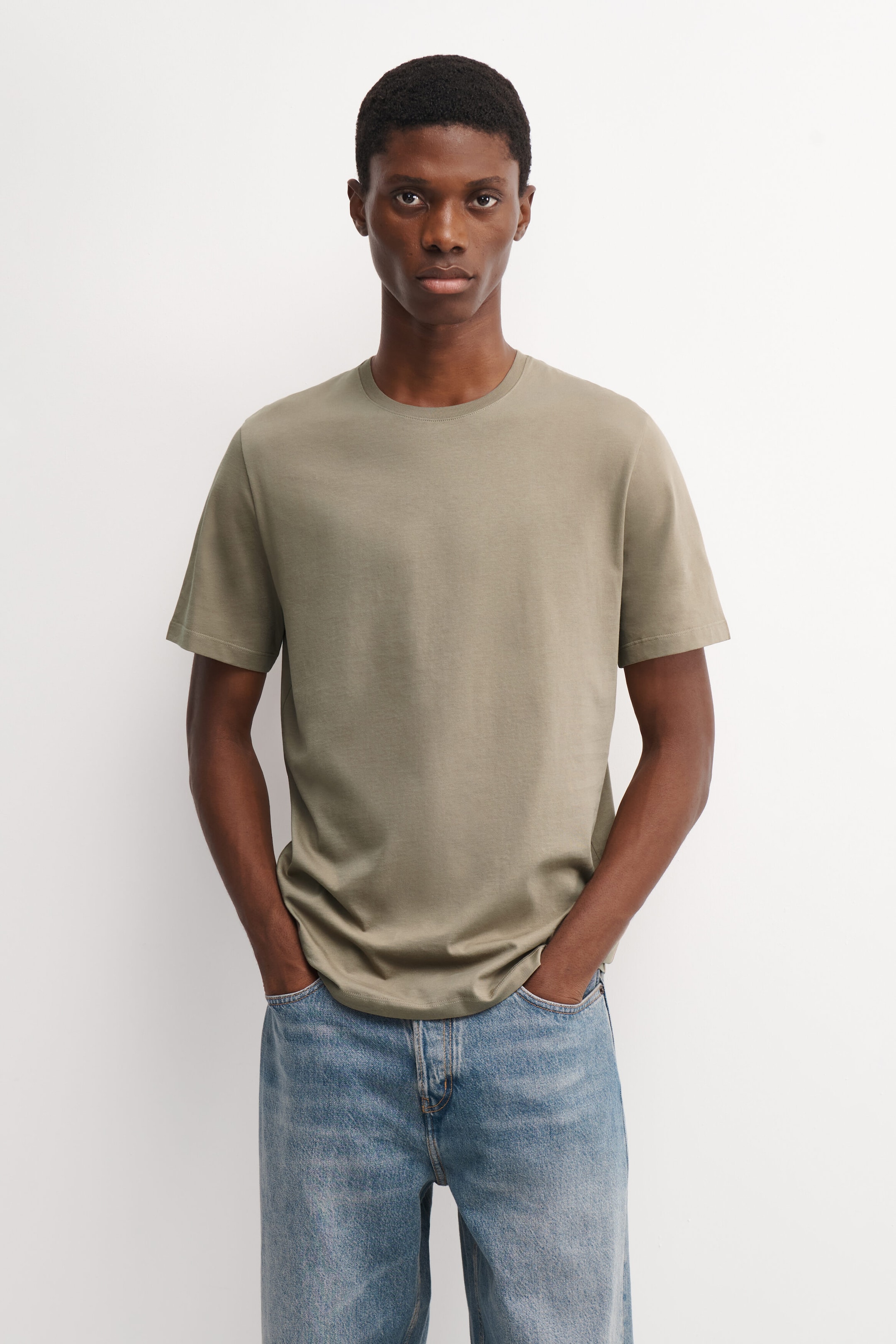 The model is wearing a light olive-green cotton crew-neck T-shirt, standing with hands tucked into the pockets of light-wash blue jeans. The short-sleeve T-shirt features smooth fabric, a classic fit, and a ribbed neckline. The model is wearing a light olive-green cotton crew-neck T-shirt, standing with hands tucked into the pockets of light-wash blue jeans. The short-sleeve T-shirt features smooth fabric, a classic fit, and a ribbed neckline.