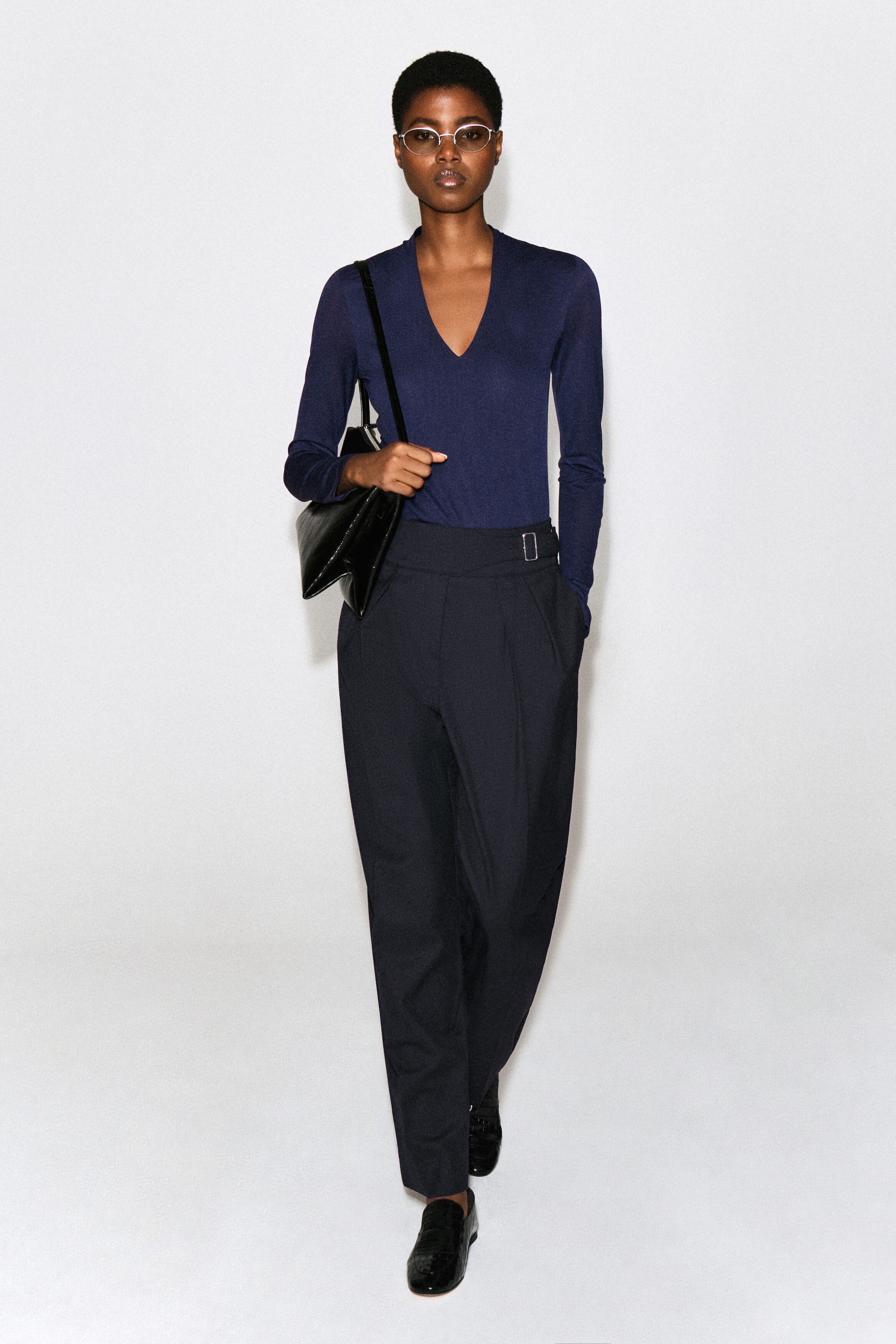 A navy blue, slim-fit, long-sleeve top with a V-neckline is worn by the model, tucked into dark, high-waisted trousers. The lightweight jersey top is styled with a black shoulder bag carried over one shoulder. A navy blue, slim-fit, long-sleeve top with a V-neckline is worn by the model, tucked into dark, high-waisted trousers. The lightweight jersey top is styled with a black shoulder bag carried over one shoulder.