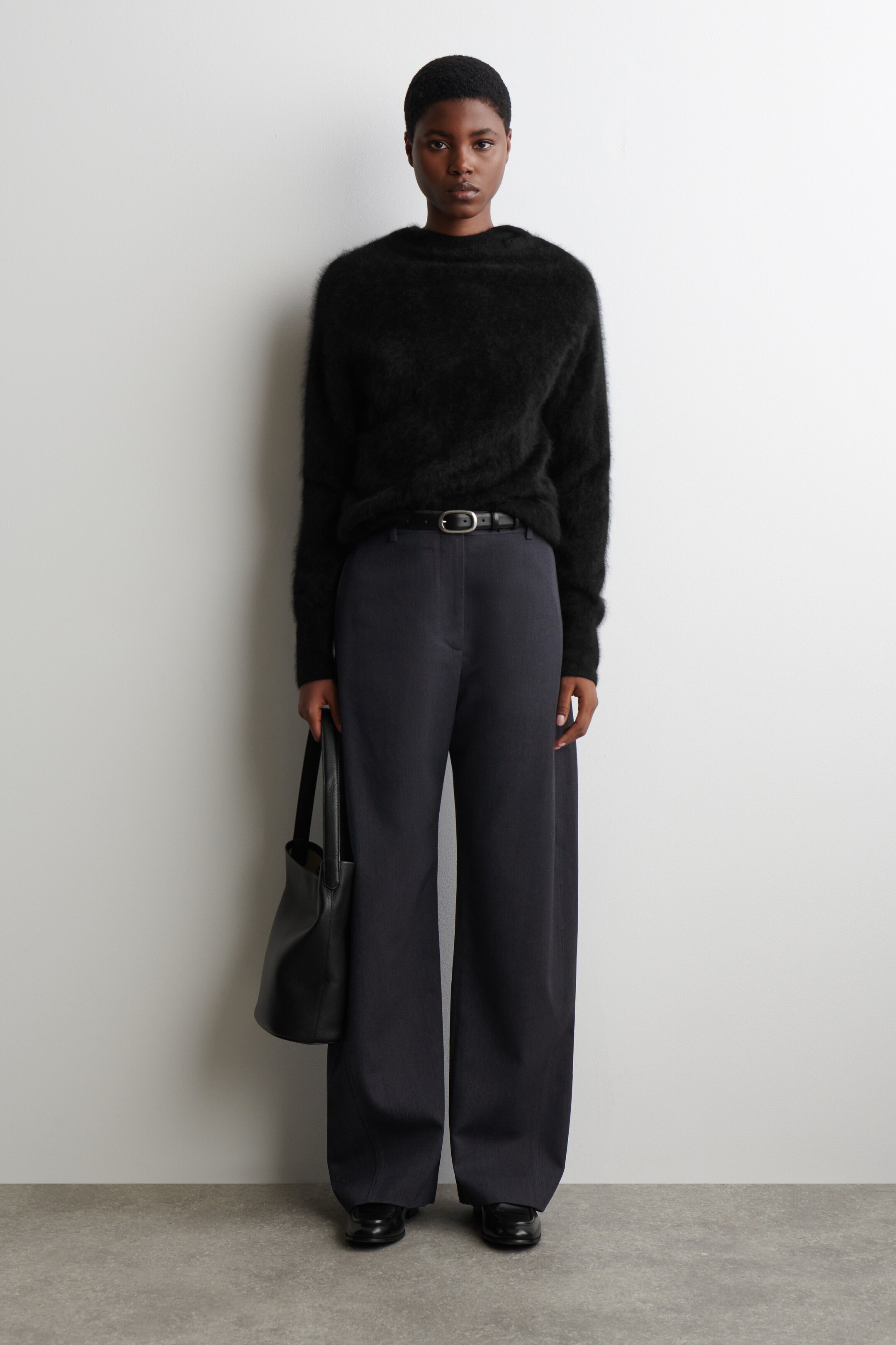 View larger image: A black fuzzy long-sleeved sweater with a high crew neck is being modeled, paired with dark charcoal wide-leg trousers featuring a visible front crease, all seen from a front-facing angle. A thin black leather belt with a silver oval buckle is cinching the waist, and a black leather tote bag is being held, completing the look with black slip-on shoes.