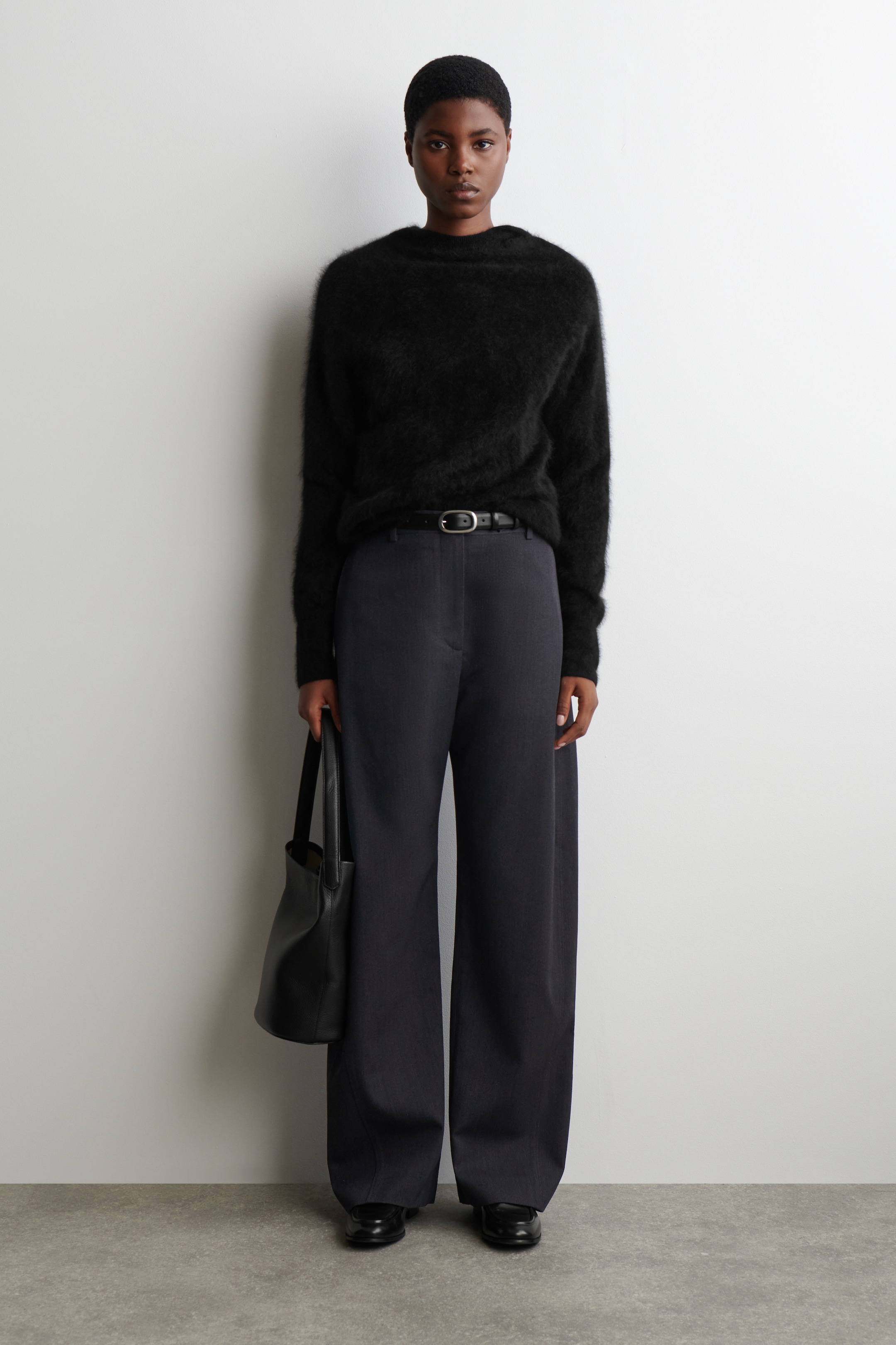 View larger image: A black fuzzy long-sleeved sweater with a high crew neck is being modeled, paired with dark charcoal wide-leg trousers featuring a visible front crease, all seen from a front-facing angle. A thin black leather belt with a silver oval buckle is cinching the waist, and a black leather tote bag is being held, completing the look with black slip-on shoes.