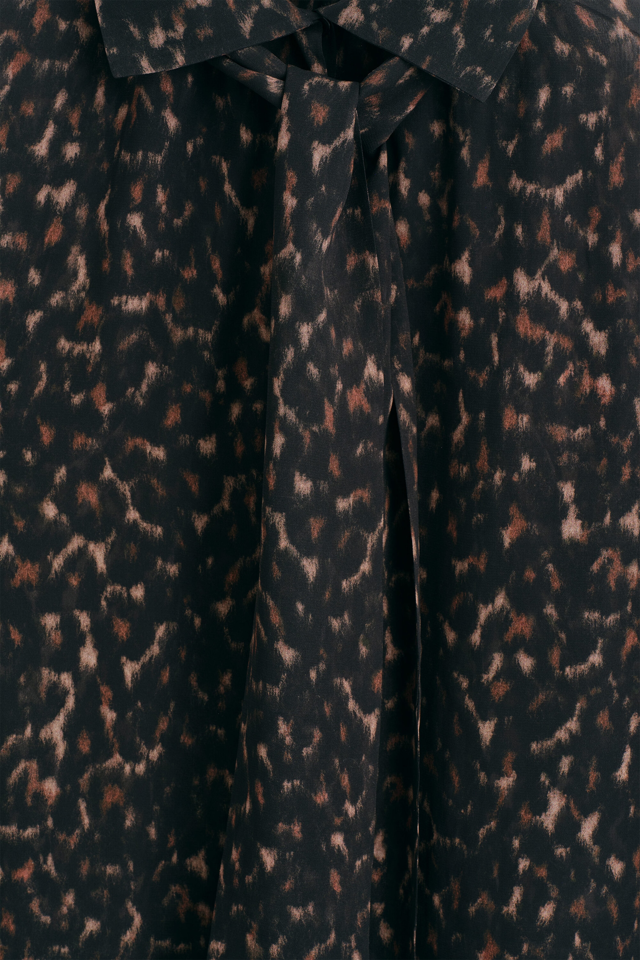 The leopard-print fluid scarf-detail top is shown in a close-up front view, highlighting its dark fabric with an abstract brown and tan leopard print. The top features a collar with an integrated scarf detail tied loosely at the centre front. The leopard-print fluid scarf-detail top is shown in a close-up front view, highlighting its dark fabric with an abstract brown and tan leopard print. The top features a collar with an integrated scarf detail tied loosely at the centre front.