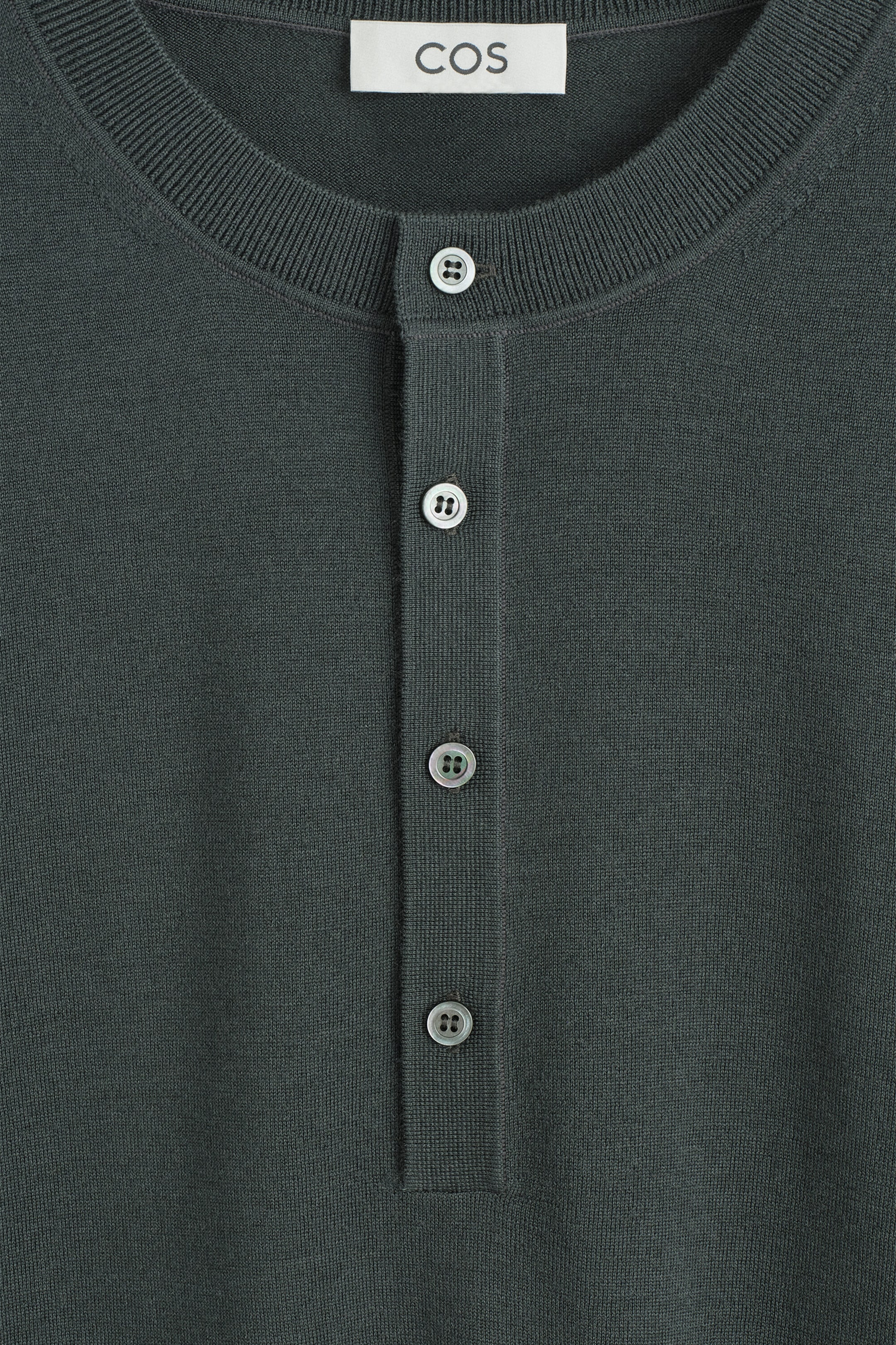A close-up front view of the Slim Merino Wool Henley Top showcasing its dark green, fine-knit fabric with a ribbed crew neckline and a vertical four-button placket.