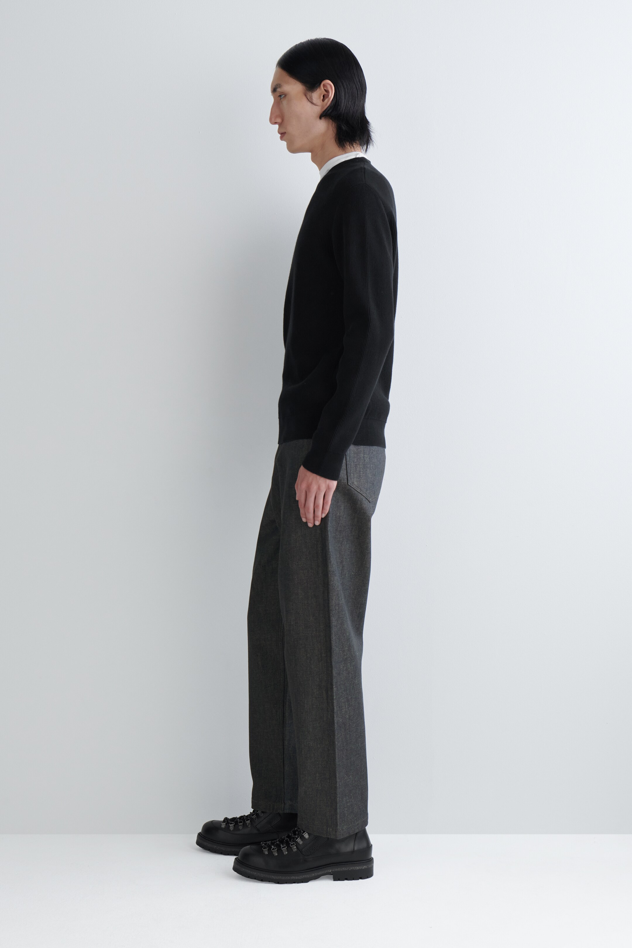 Agrandir l'image: Un pull en tricot noir avec des côtes verticales subtiles et des poignets côtelés est présenté de profil, superposé à un col intérieur blanc, et associé à un pantalon gris foncé à coupe décontractée et jambes droites, ainsi qu'à des bottes noires à lacets avec une semelle épaisse.