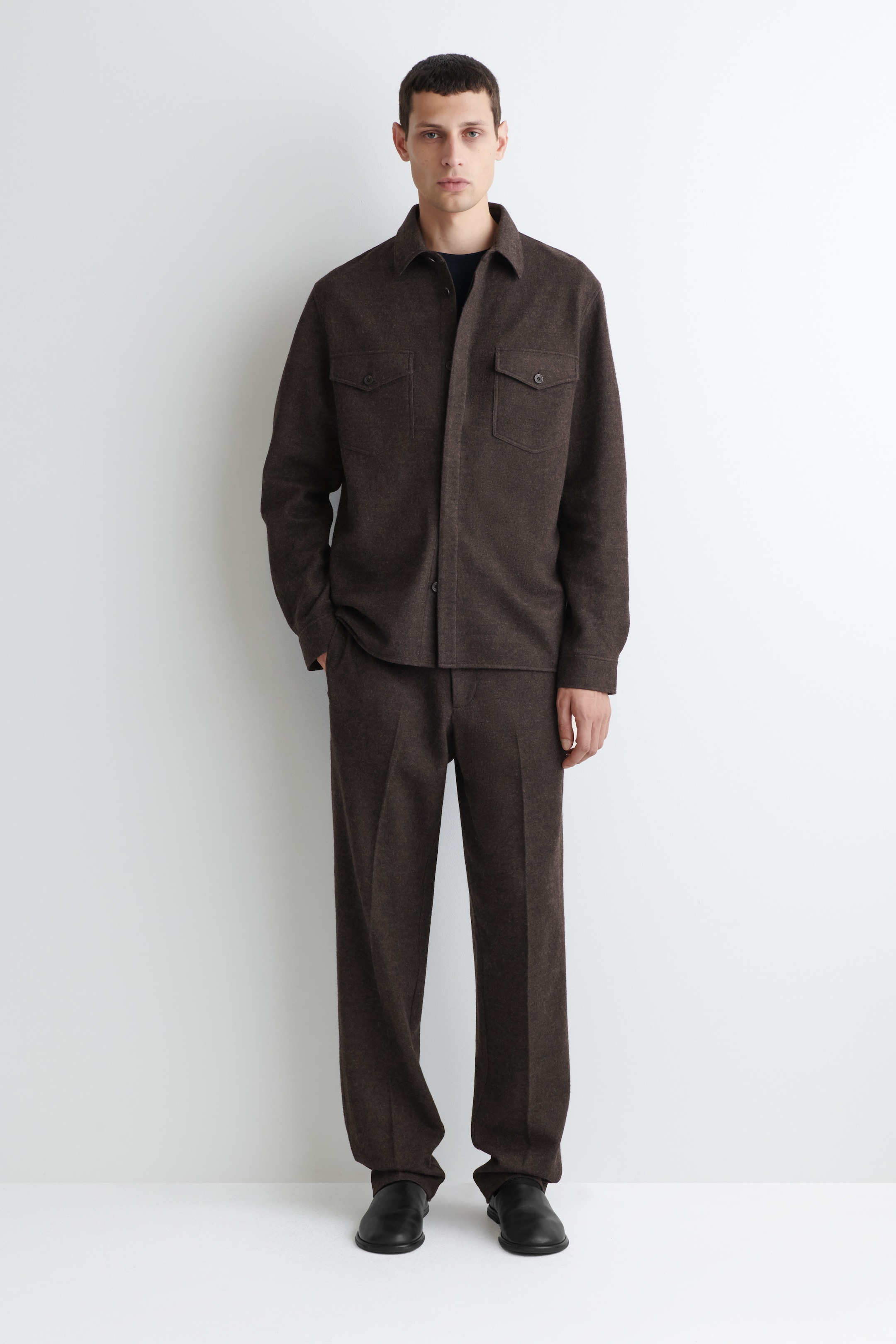 Agrandir l'image: Une surchemise chinée brun foncé et un pantalon droit assorti sont présentés comme une tenue complète. La surchemise présente un col classique, une fermeture à boutons sur le devant et deux poches plaquées boutonnées sur la poitrine, et est portée par-dessus un t-shirt à col rond foncé. Le pantalon complète la coupe décontractée et est associé à des chaussures plates noires.