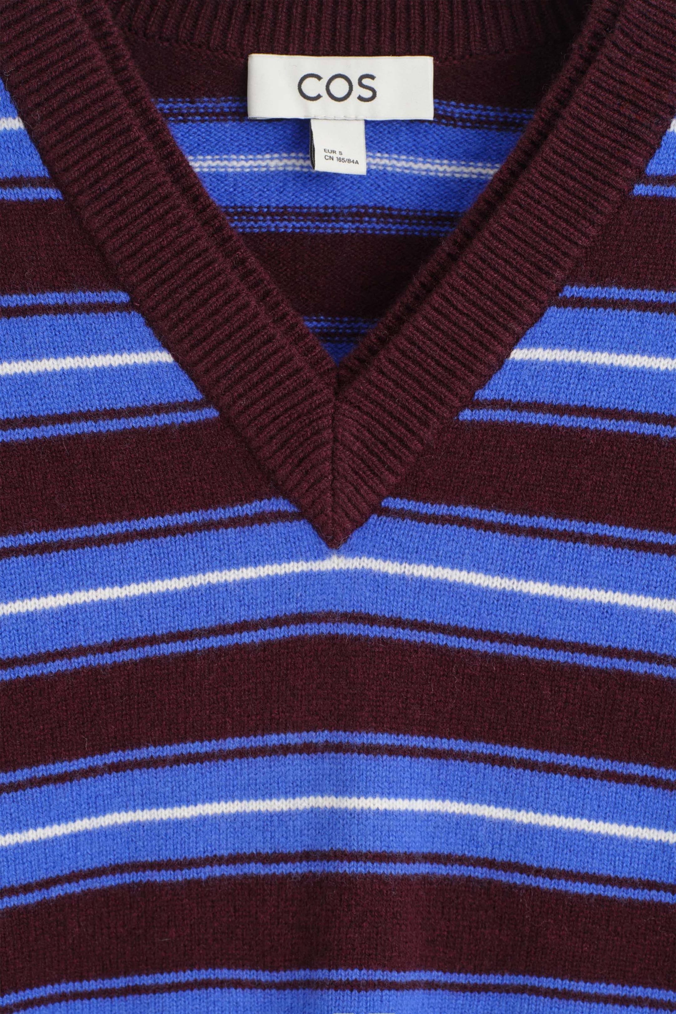 The Merino Wool Layered V-Neck Sweater is shown in a close-up front view, highlighting its ribbed burgundy V-neck collar and horizontal striped pattern. The design combines burgundy, bright blue, and thin white stripes across a fine-knit fabric, with a brand label visible at the inner neckline. The Merino Wool Layered V-Neck Sweater is shown in a close-up front view, highlighting its ribbed burgundy V-neck collar and horizontal striped pattern. The design combines burgundy, bright blue, and thin white stripes across a fine-knit fabric, with a brand label visible at the inner neckline.