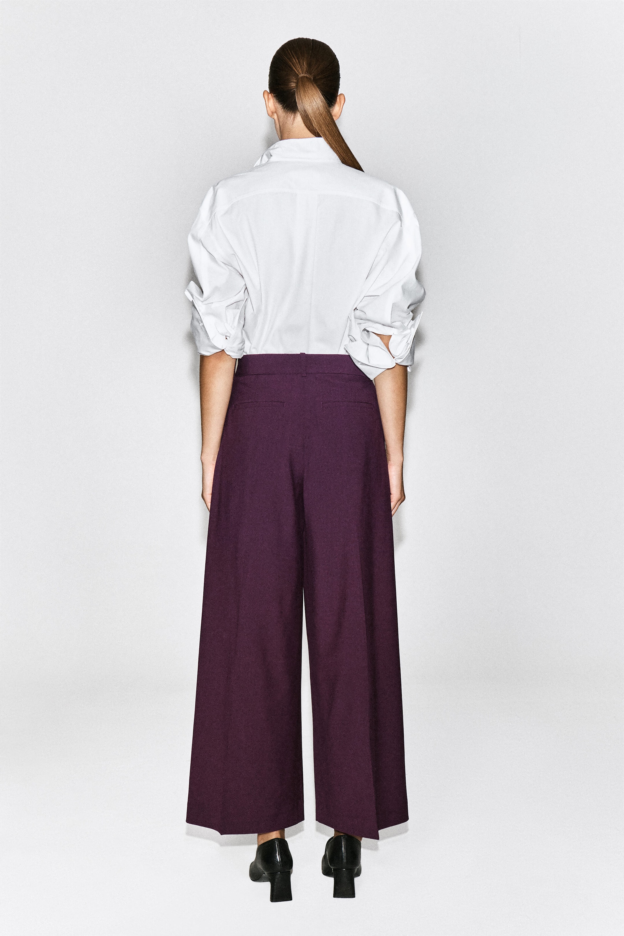 I pantaloni a gamba larga color prugna sono indossati dalla modella, mostrata di spalle. I pantaloni a vita alta presentano tasche a filetto sul retro e pieghe centrali stirate, abbinati a una camicia bianca infilata nei pantaloni e a scarpe nere con tacco. I pantaloni a gamba larga color prugna sono indossati dalla modella, mostrata di spalle. I pantaloni a vita alta presentano tasche a filetto sul retro e pieghe centrali stirate, abbinati a una camicia bianca infilata nei pantaloni e a scarpe nere con tacco.