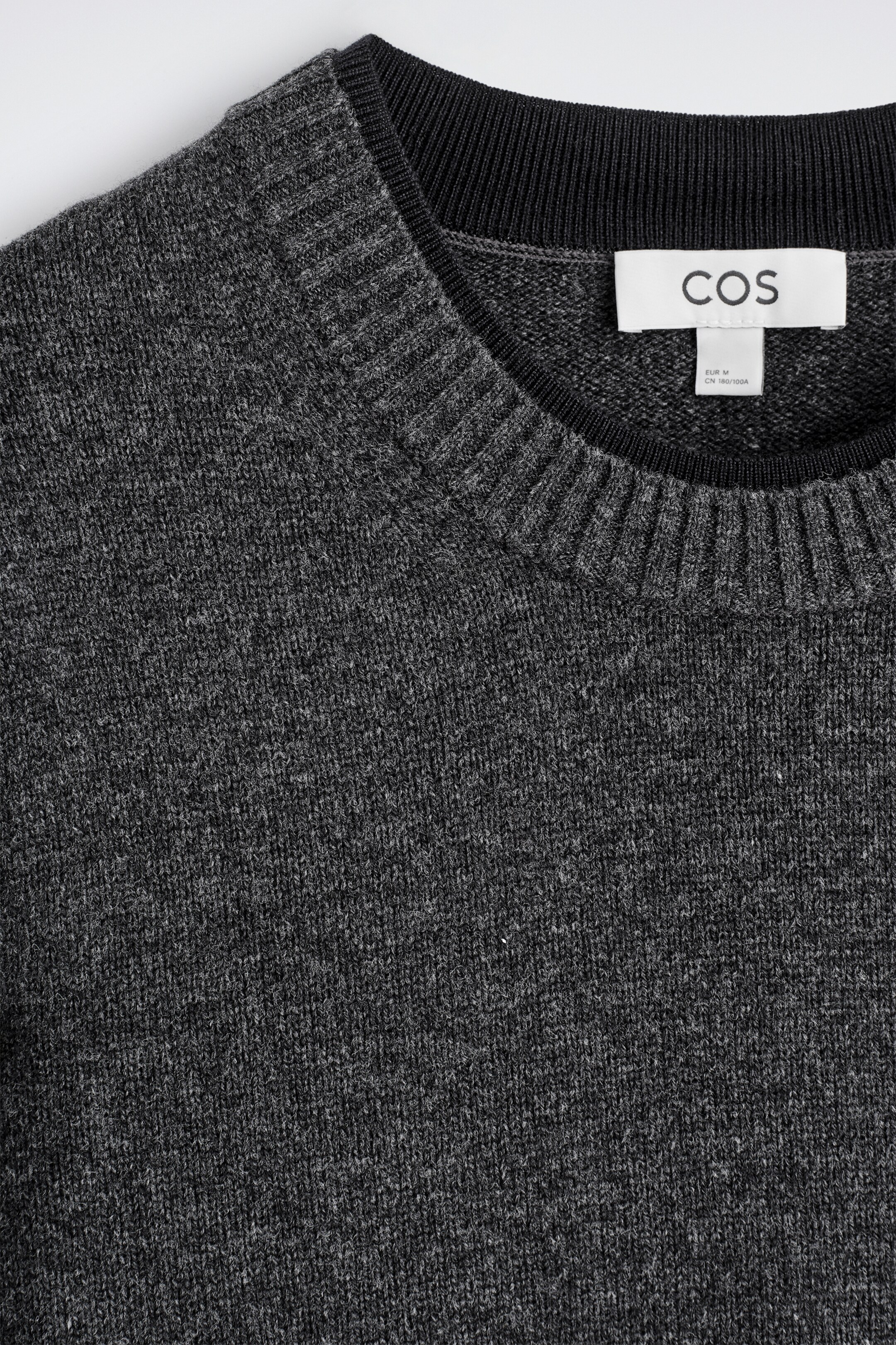 View larger image: A charcoal grey merino wool jumper is being shown in a close-up of its upper front. It is featuring a crew neck with a ribbed knit collar, beneath which a black inner trim is visible, alongside a white COS brand tag sewn into the neckline.
