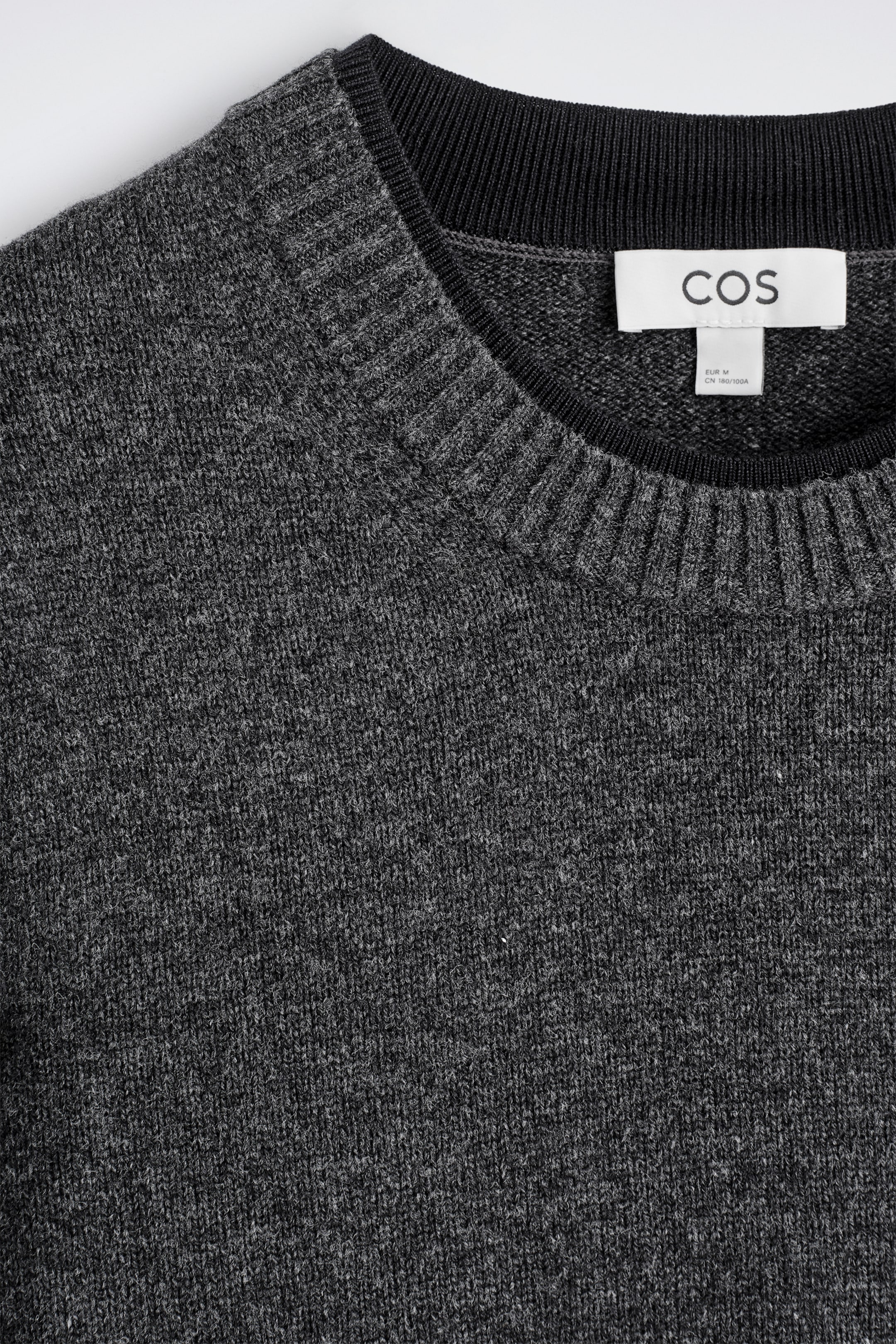 Grösseres Bild ansehen: Ein anthrazitgrauer Merinowollpullover wird in einer Nahaufnahme seiner oberen Vorderseite gezeigt. Er verfügt über einen Rundhalsausschnitt mit einem gerippten Strickkragen, unter dem ein schwarzer Innensaum sichtbar ist, neben einem weißen COS-Markenetikett, das in den Halsausschnitt eingenäht ist.