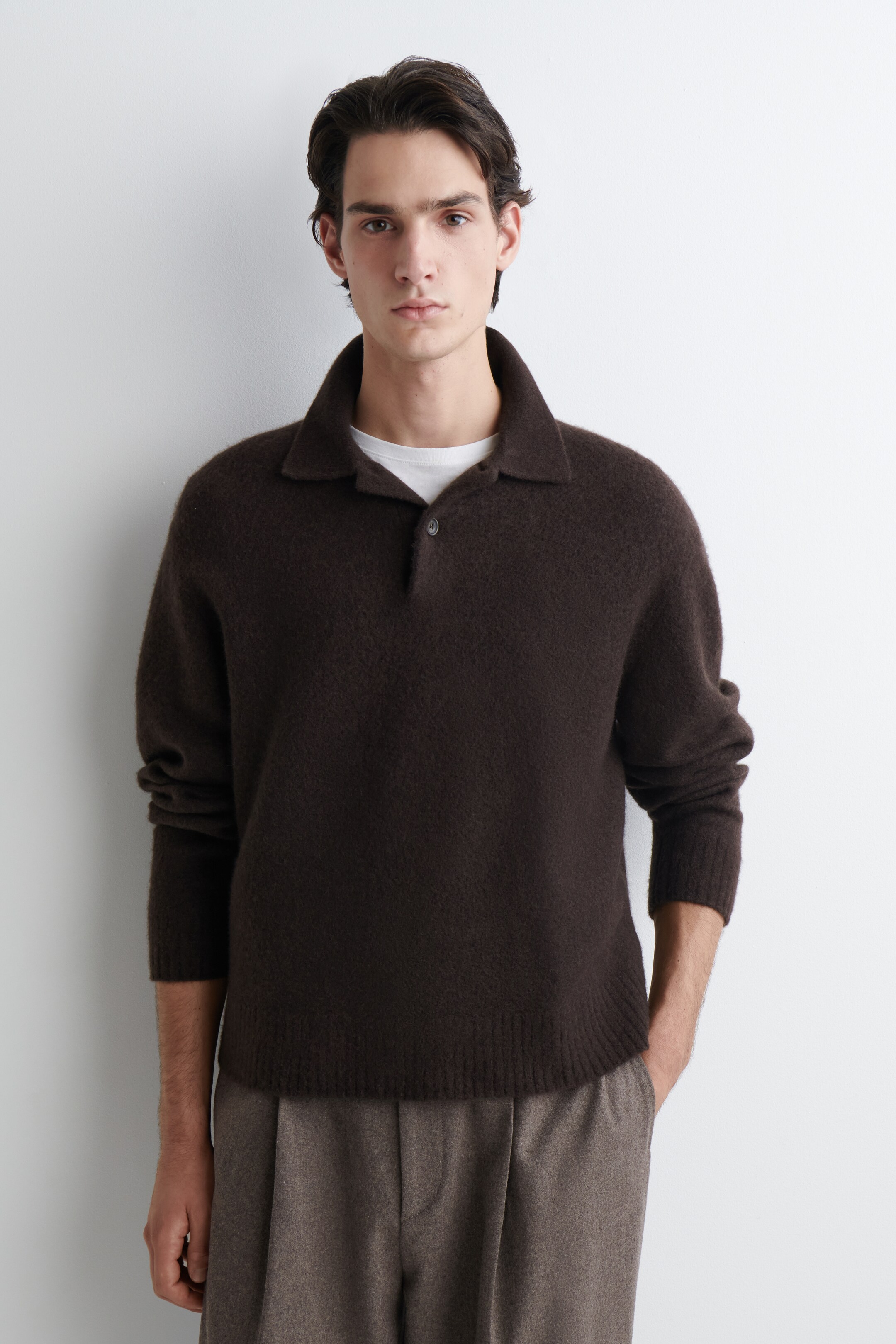 Agrandir l'image: Un pull en maille polo marron foncé est présenté de face, avec une texture duveteuse, un col polo et une patte à un bouton. Il est stylé avec un t-shirt blanc visible sous le col et un pantalon plissé marron clair visible sous son ourlet et ses poignets côtelés.