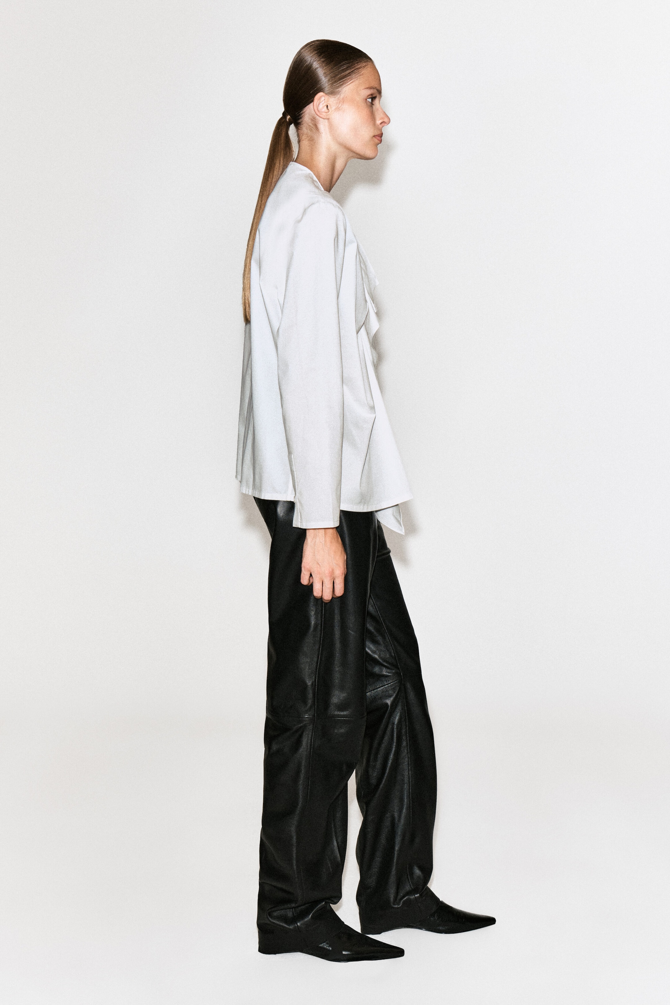 A white ruffled cotton blouse with long sleeves and a relaxed silhouette is being worn by the model standing in side profile. The blouse is styled with black leather pants and black pointed-toe shoes.