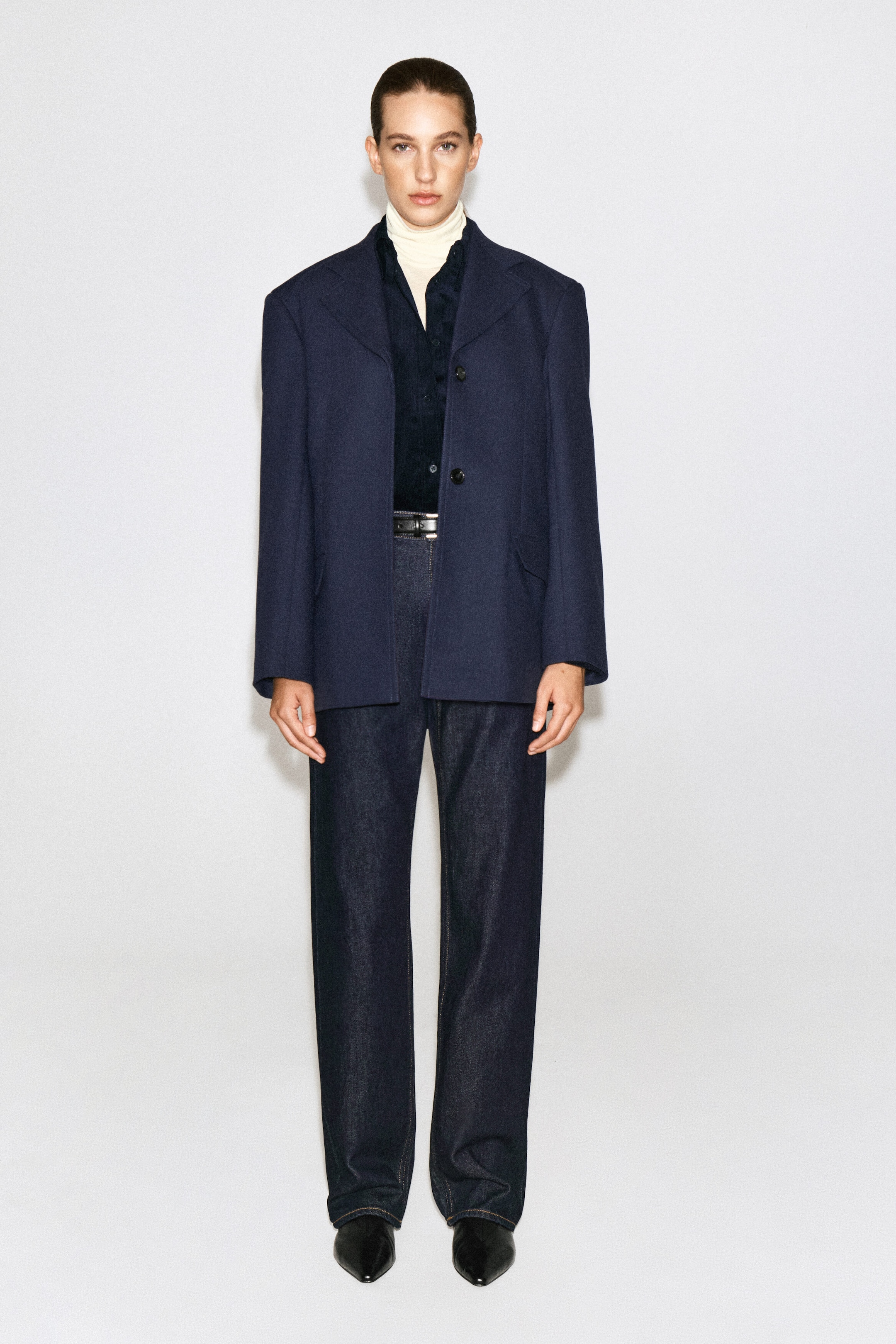 The model is wearing a navy cotton corduroy shirt layered over a cream turtleneck and styled under an open, structured navy blazer. The buttoned-up shirt is tucked into dark-wash denim jeans fastened with a black belt. The model is wearing a navy cotton corduroy shirt layered over a cream turtleneck and styled under an open, structured navy blazer. The buttoned-up shirt is tucked into dark-wash denim jeans fastened with a black belt.