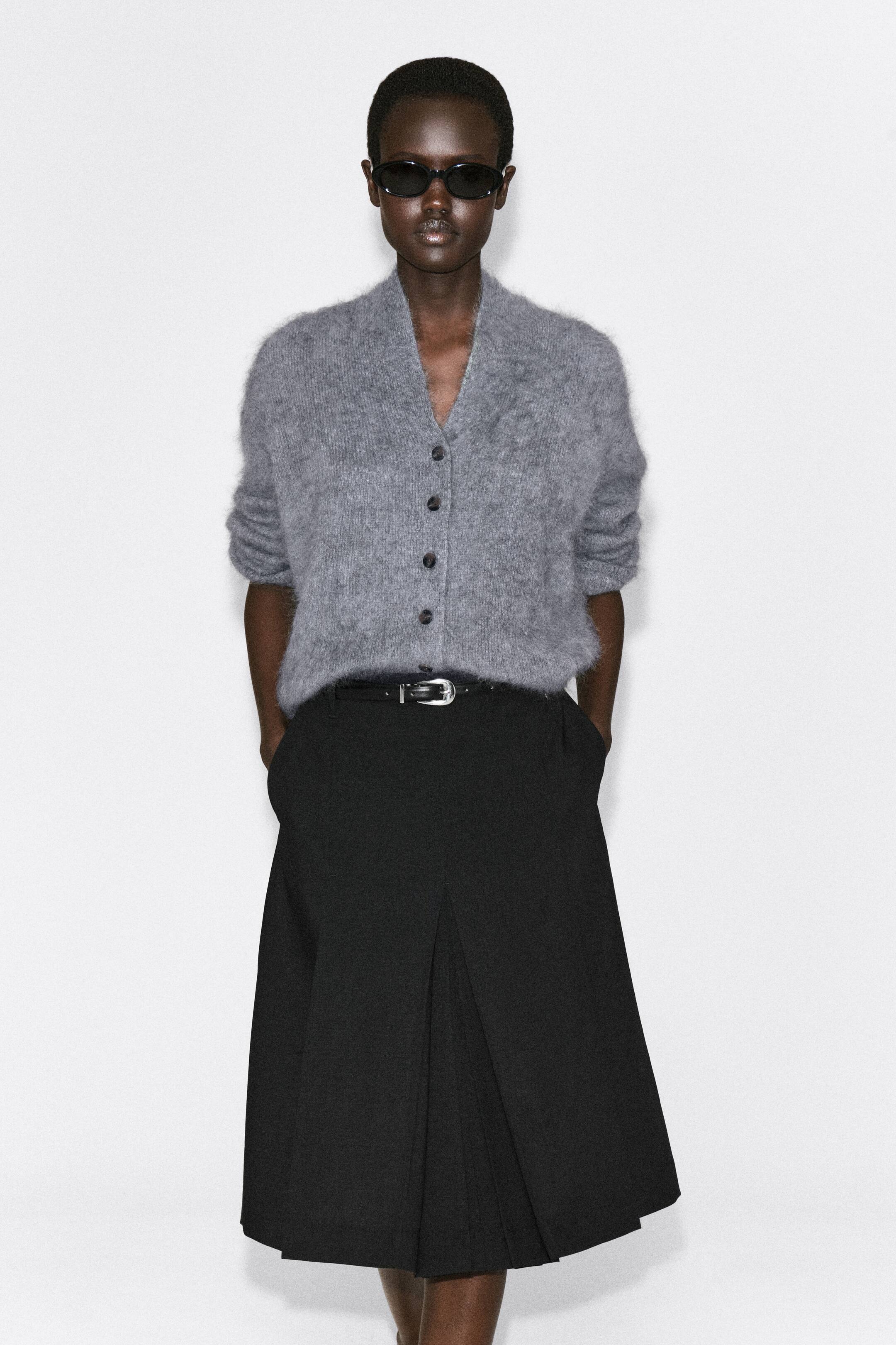 Agrandir l'image: Ce cardigan gris en laine mérinos brossée drapée est présenté de face, avec un col en V, une fermeture boutonnée avec des boutons foncés et une finition duveteuse texturée. Le cardigan est stylisé rentré dans une jupe noire ceinturée, avec ses longues manches retroussées jusqu’aux coudes pour créer une silhouette décontractée.