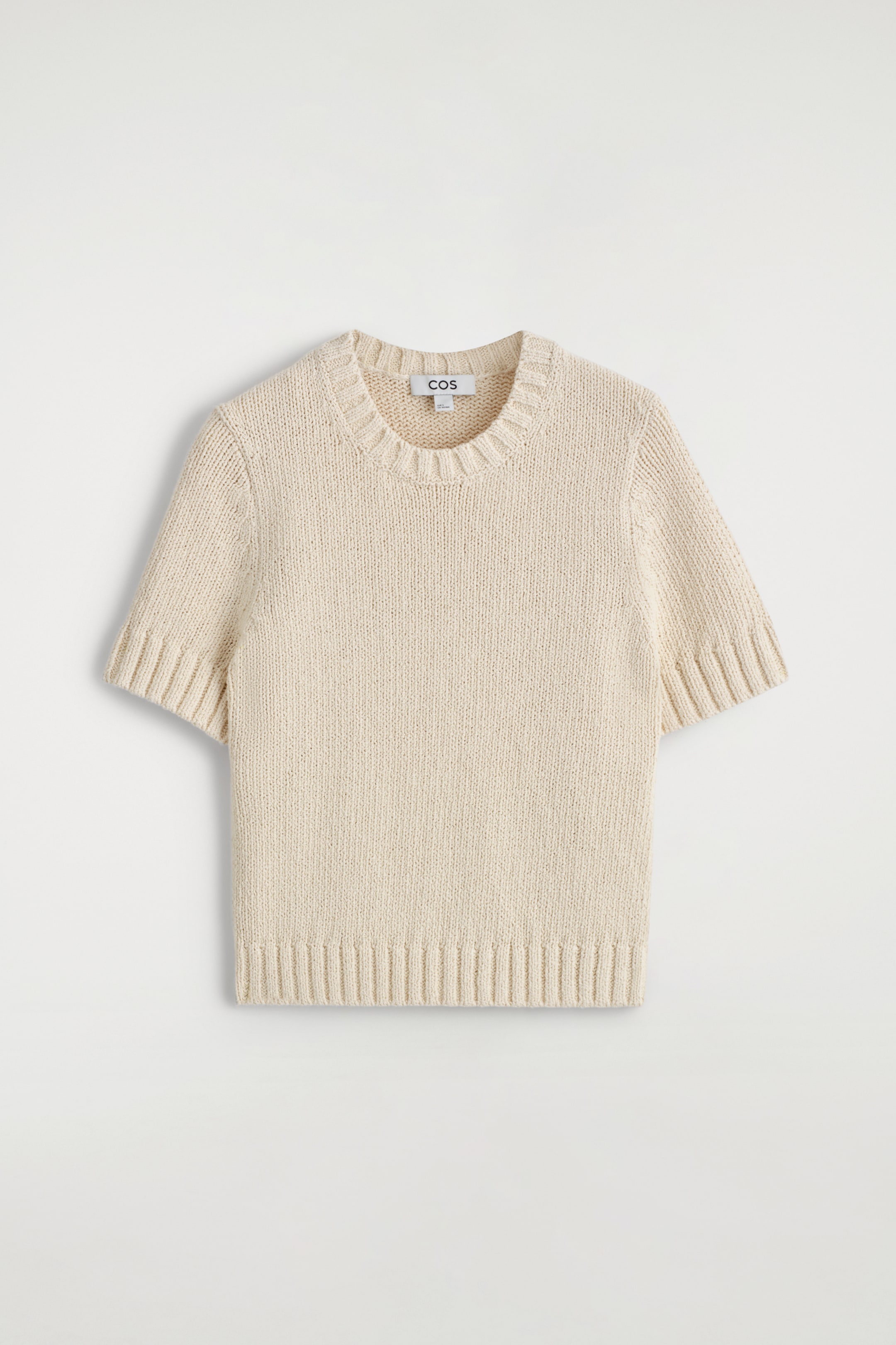 Grösseres Bild ansehen: Ein cremefarbenes, kurzärmeliges Strickoberteil liegt flach und zeigt seine Vorderseite. Es präsentiert einen schlichten Strickkörper mit einem gerippten Rundhalsausschnitt, gerippten Bündchen an den kurzen Ärmeln und einem gerippten Saum. Ein 'COS'-Etikett ist im Halsausschnitt sichtbar.