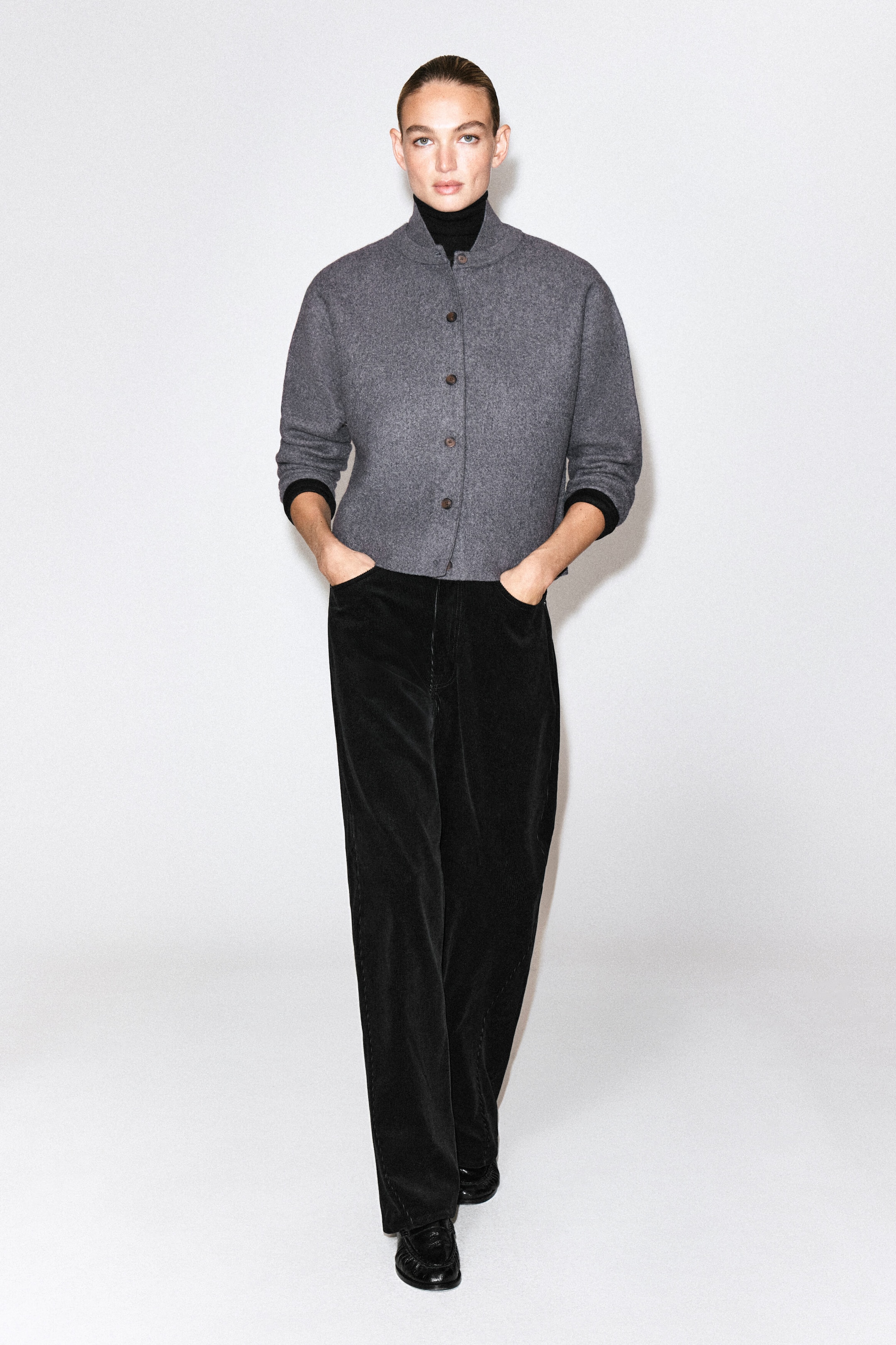 A gray boiled wool cardigan with a high collar and front buttons is being worn by the model over a black turtleneck. The model is standing with their hands in the pockets of black wide-leg trousers. A gray boiled wool cardigan with a high collar and front buttons is being worn by the model over a black turtleneck. The model is standing with their hands in the pockets of black wide-leg trousers.