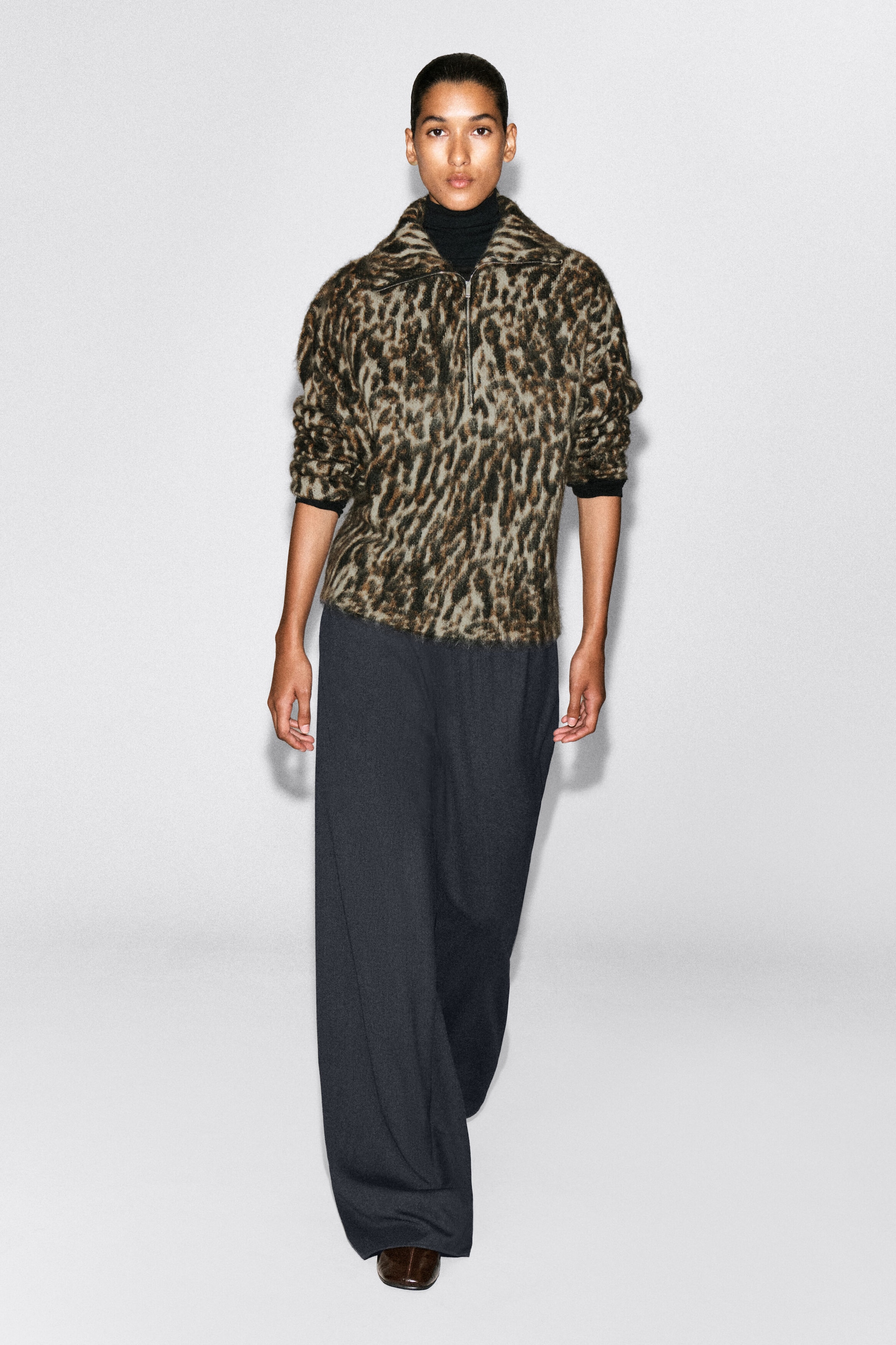 A leopard jacquard jumper with a fuzzy, brushed texture is shown from the front on a model. The jumper features a zip-up funnel neck, worn partially unzipped over a black roll neck, and is styled with dark grey wide-leg trousers. A leopard jacquard jumper with a fuzzy, brushed texture is shown from the front on a model. The jumper features a zip-up funnel neck, worn partially unzipped over a black roll neck, and is styled with dark grey wide-leg trousers.