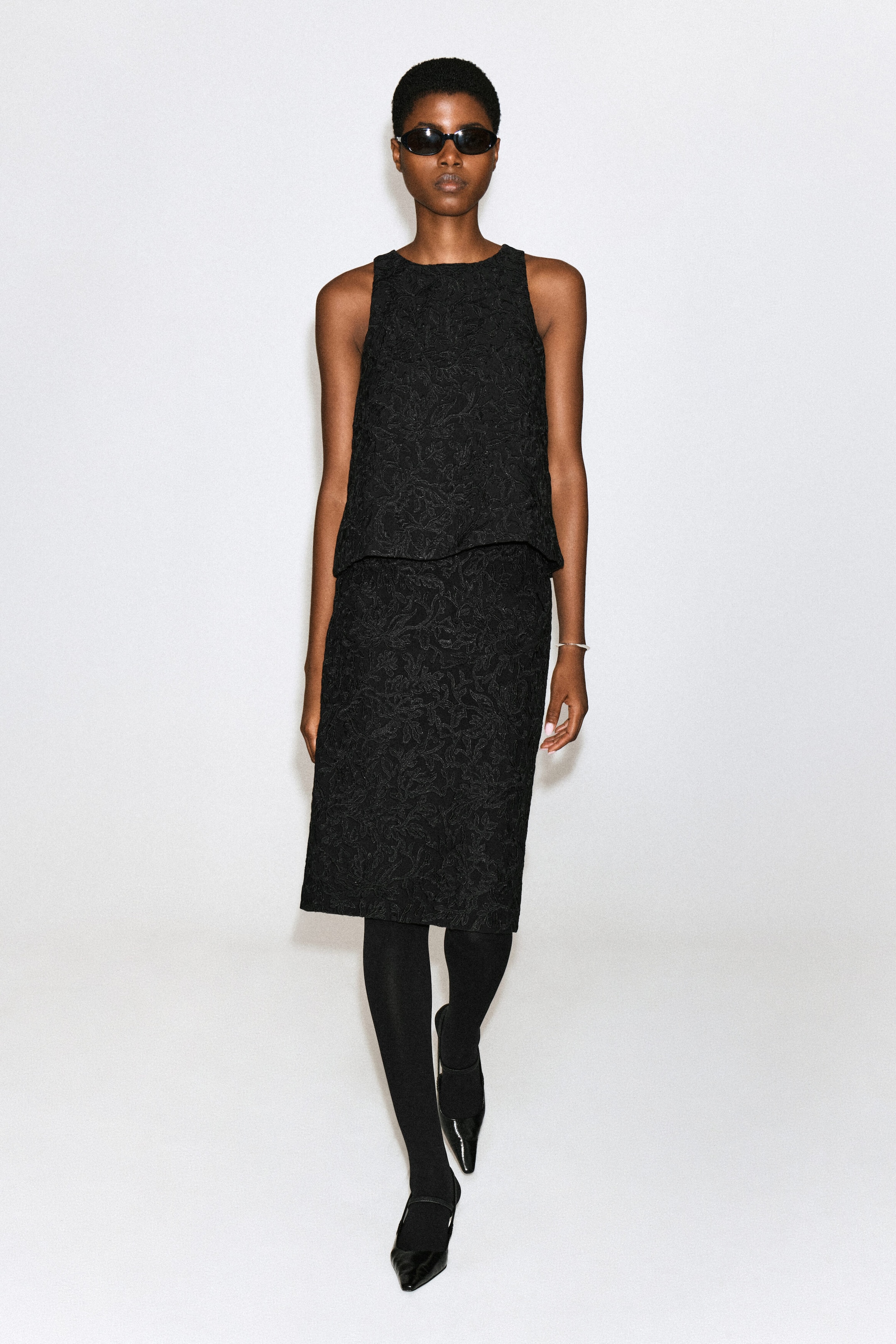 A black midi skirt featuring a textured floral lace pattern is worn by the model. The skirt is styled with a matching sleeveless top, black tights, and black pointed-toe heels. A black midi skirt featuring a textured floral lace pattern is worn by the model. The skirt is styled with a matching sleeveless top, black tights, and black pointed-toe heels.