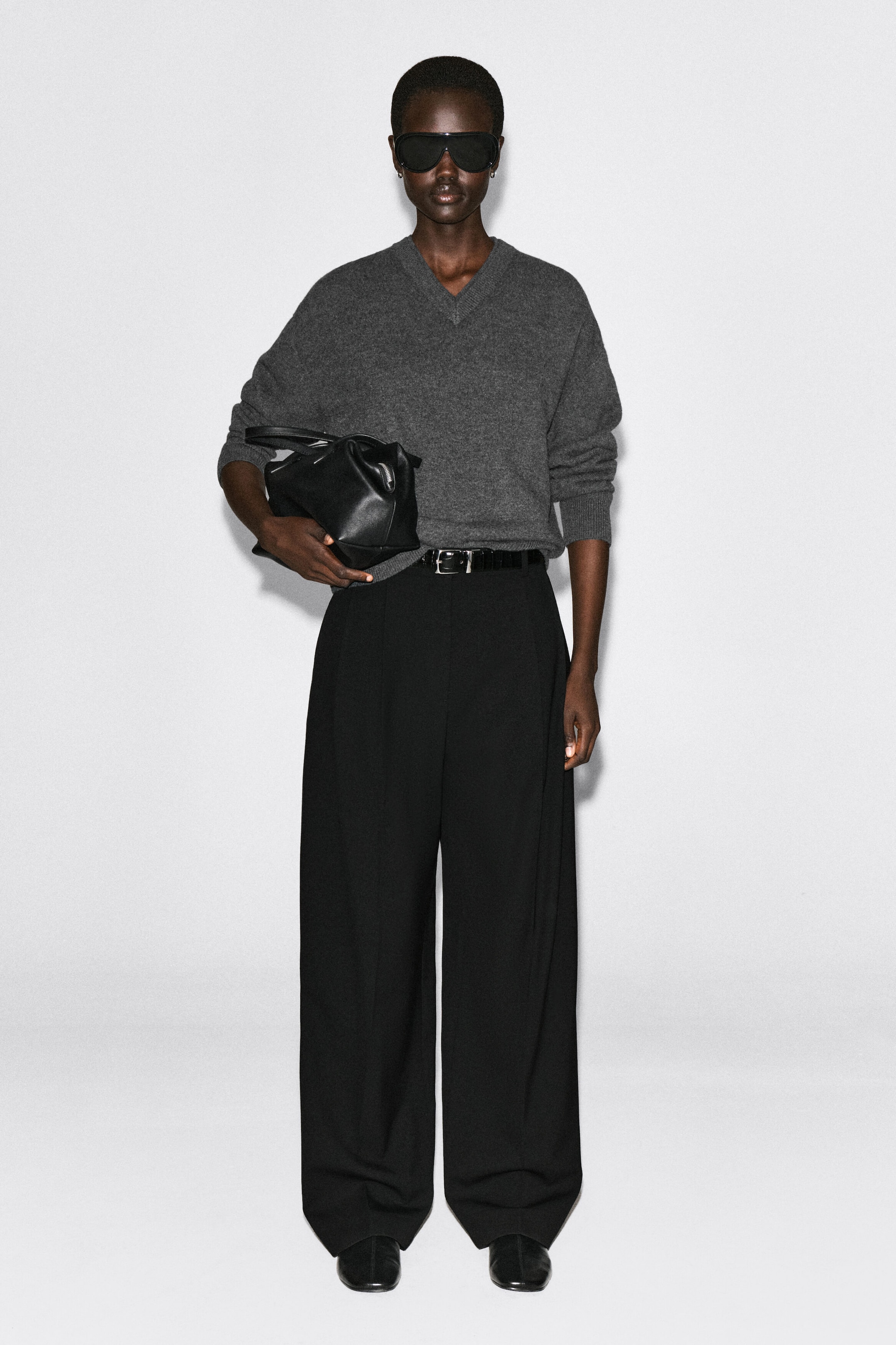 The front-facing merino wool layered V-neck sweater features a relaxed fit in a grey mélange knit with a distinctive double-layered V-neckline. The sweater is shown with long sleeves and ribbed cuffs, and is partially tucked into wide-leg black trousers to showcase its soft drape. The front-facing merino wool layered V-neck sweater features a relaxed fit in a grey mélange knit with a distinctive double-layered V-neckline. The sweater is shown with long sleeves and ribbed cuffs, and is partially tucked into wide-leg black trousers to showcase its soft drape.
