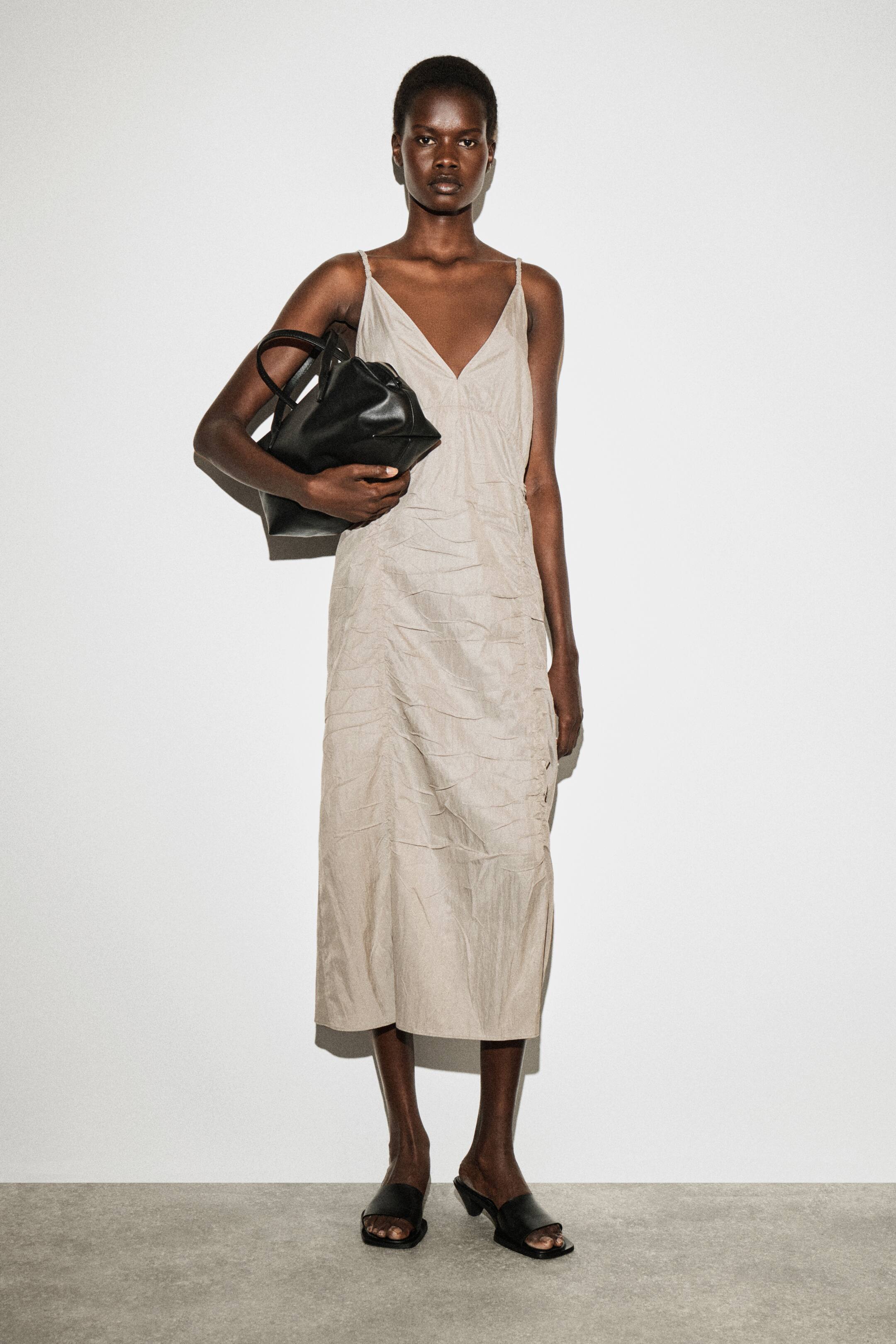 הצגת תמונה גדולה יותר: שמלת סליפ בצבע בז' בהיר עם מחשוף V עמוק ורצועות ספגטי עדינות נלבשת על ידי הדוגמנית. השמלה כוללת כיווצים לאורך הצדדים, והדוגמנית מחזיקה תיק יד שחור ונועלת סנדלים שחורים.