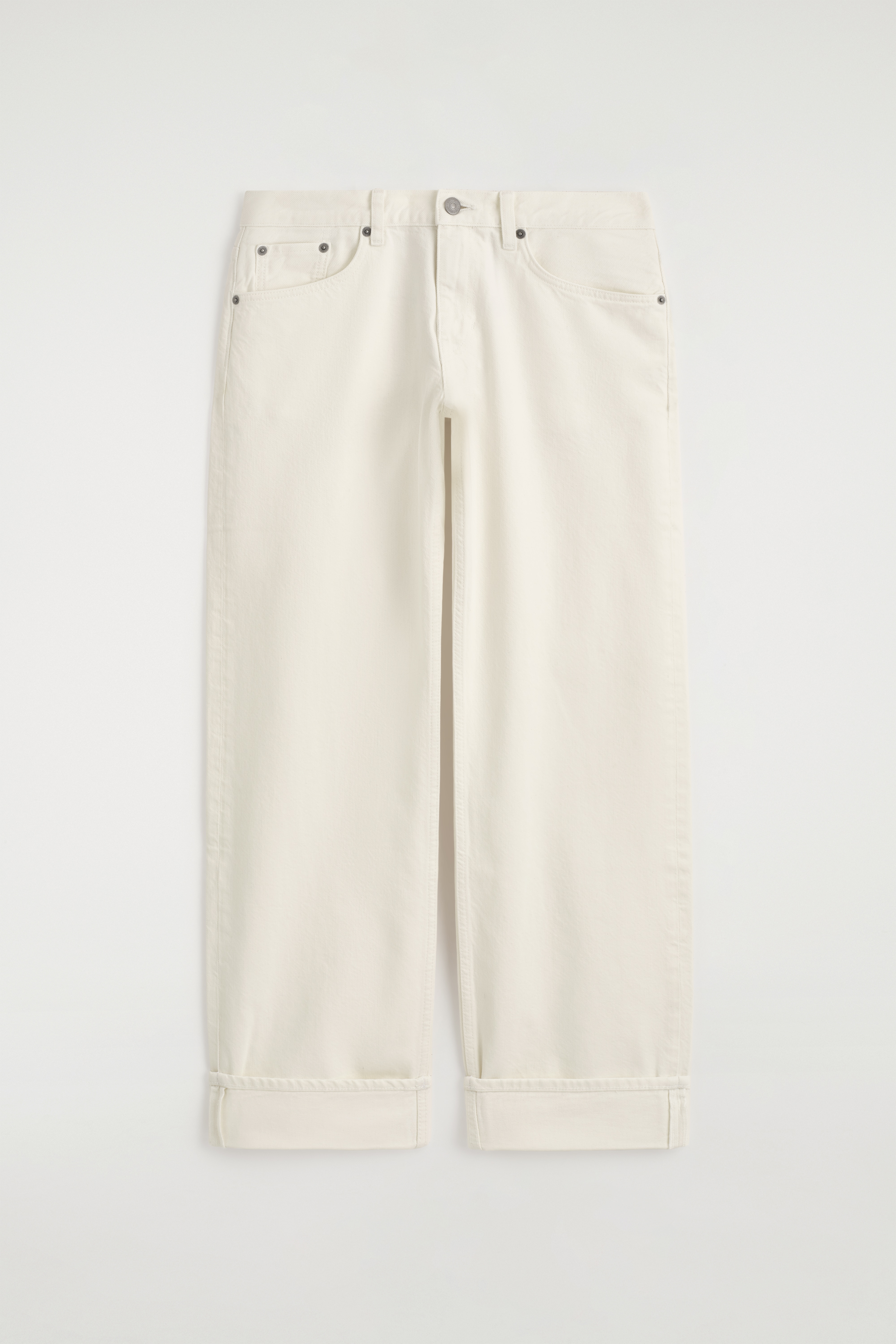 De Folde jeans met gebogen pijpen en omgeslagen zoom in off-white denim worden gedragen door het model, dat staat met Ã©Ã©n hand rustend in de voorzak. De jeans heeft een hoge taille, een losse pasvorm met gebogen pijpen en brede omgeslagen boorden, hier gestyled met een zwarte riem en een zwart leren hemd.