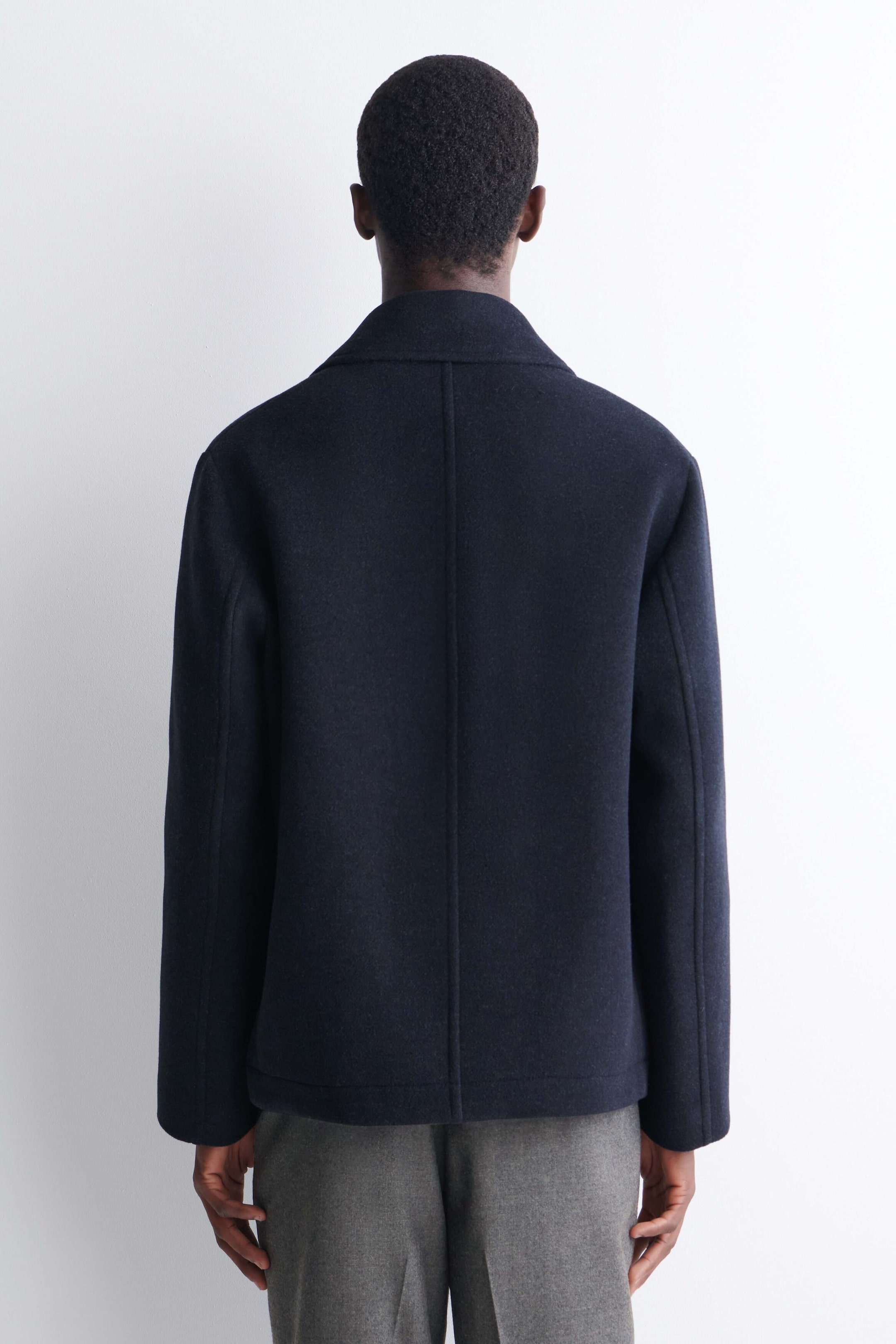 The model is wearing a dark navy double-breasted wool short pea coat, shown from the back. The coat features a structured collar, a central back seam and long sleeves, styled with grey trousers.