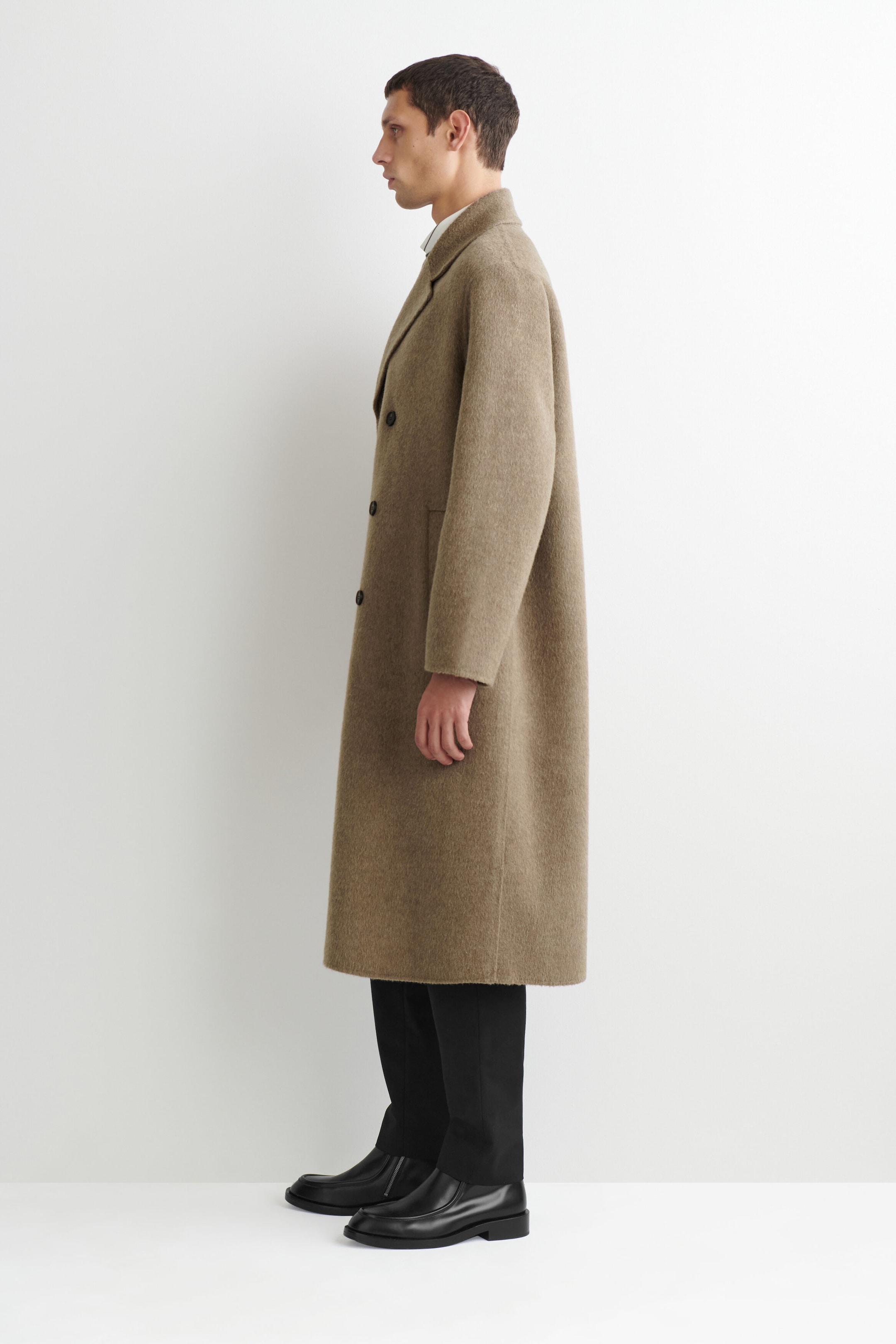 The double-faced wool-blend long coat in a textured taupe-brown drapes in a straight, calf-length silhouette, shown from a left-facing profile view. The coat features structured notch lapels and a double-breasted button design, layered over black trousers and black boots to highlight its clean lines and relaxed fit.