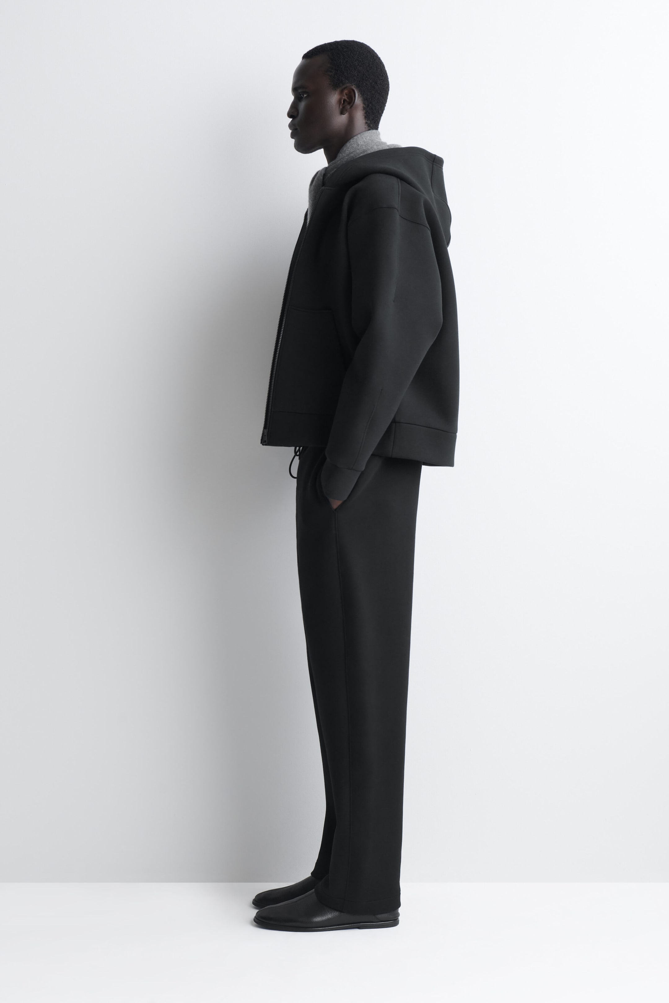A black matching set, shown in left-facing side profile, features a structured, oversized zip-up hoodie with dropped shoulders and a large hood, paired with matching straight-leg trousers. The jacket is layered over a gray knit collar, and the trousers fall straight over black leather slip-on shoes.